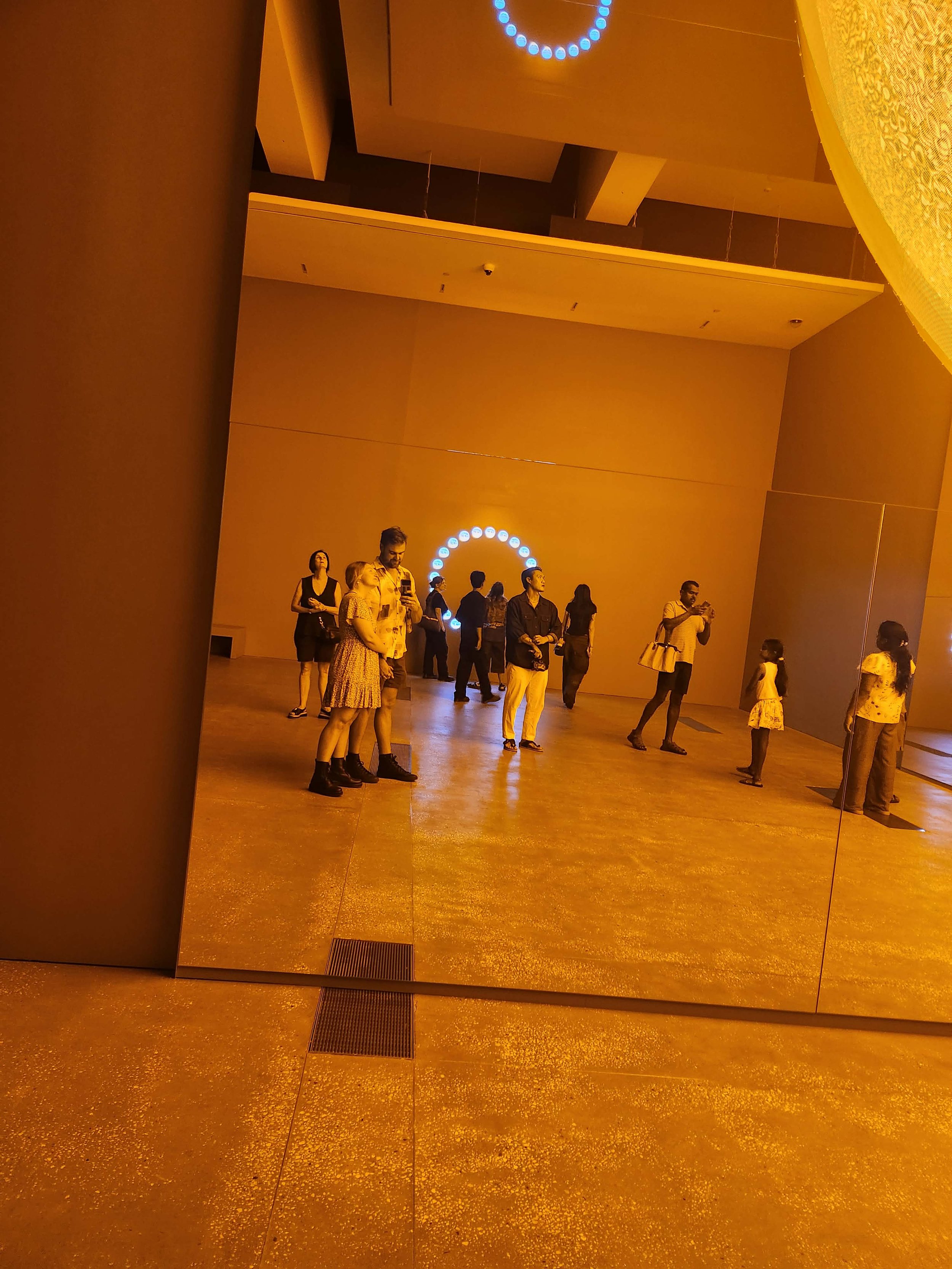 People standing and walking in a spacious, well-lit interior with golden flooring, large beige walls, and a decorative blue circular light fixture on the ceiling.
