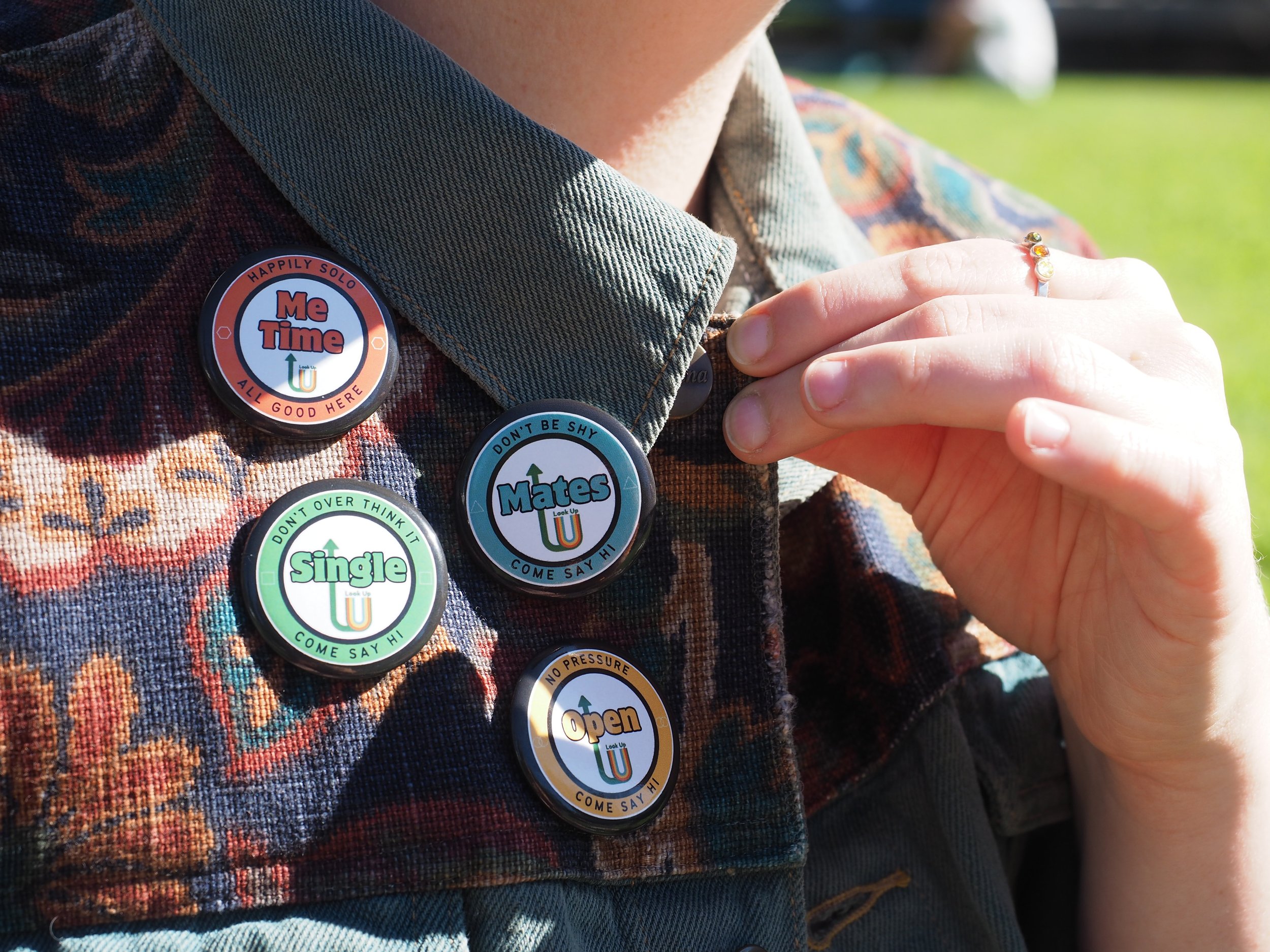 Close-up of a person wearing a denim jacket with colorful patches and buttons, including one that says 'Me Time' and another that says 'Yates,' and some other buttons with various messages, outdoors with green grass in the background.