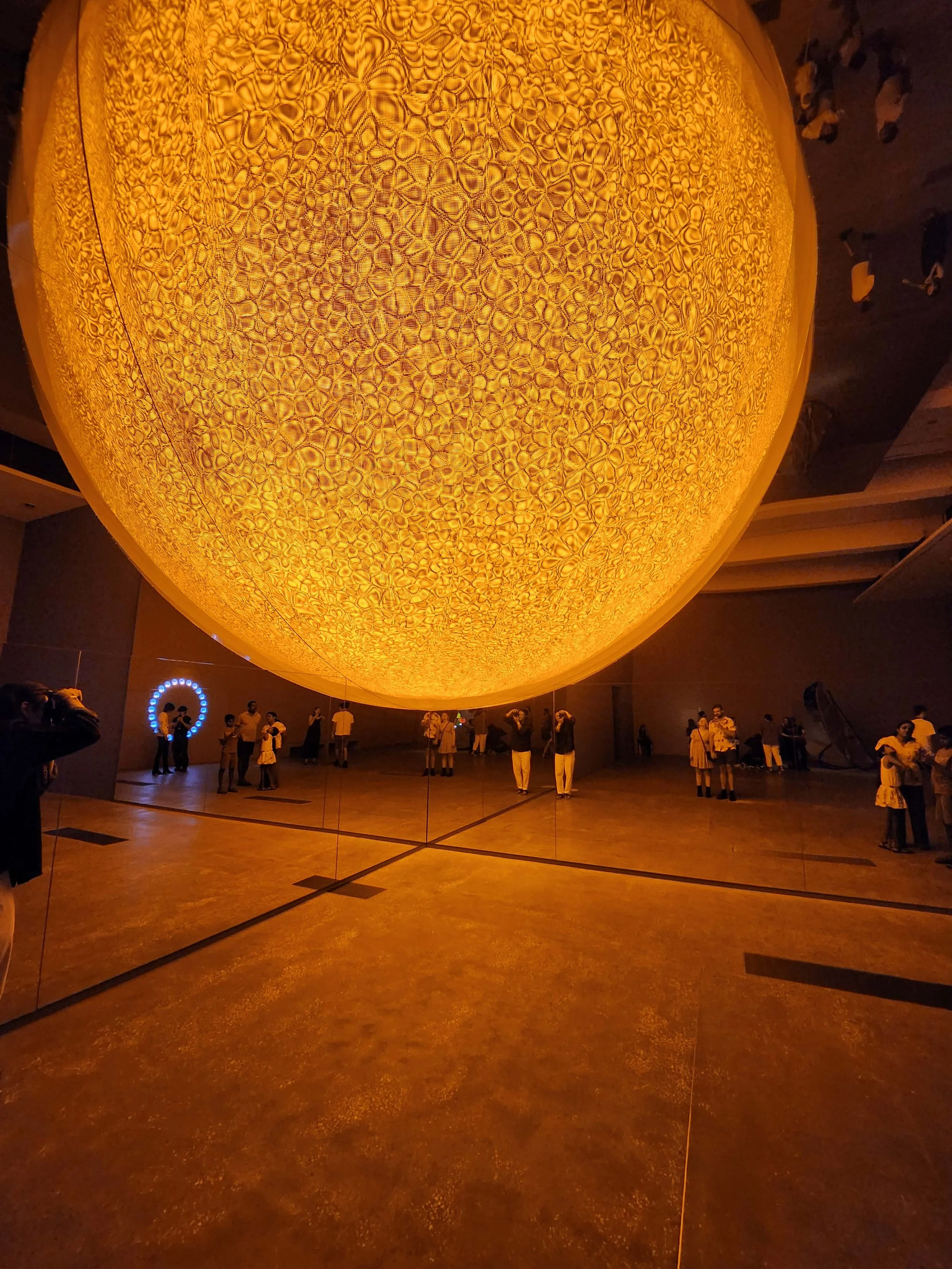 People observing an illuminated, textured, large, yellow, dome-shaped art installation in a dark room.