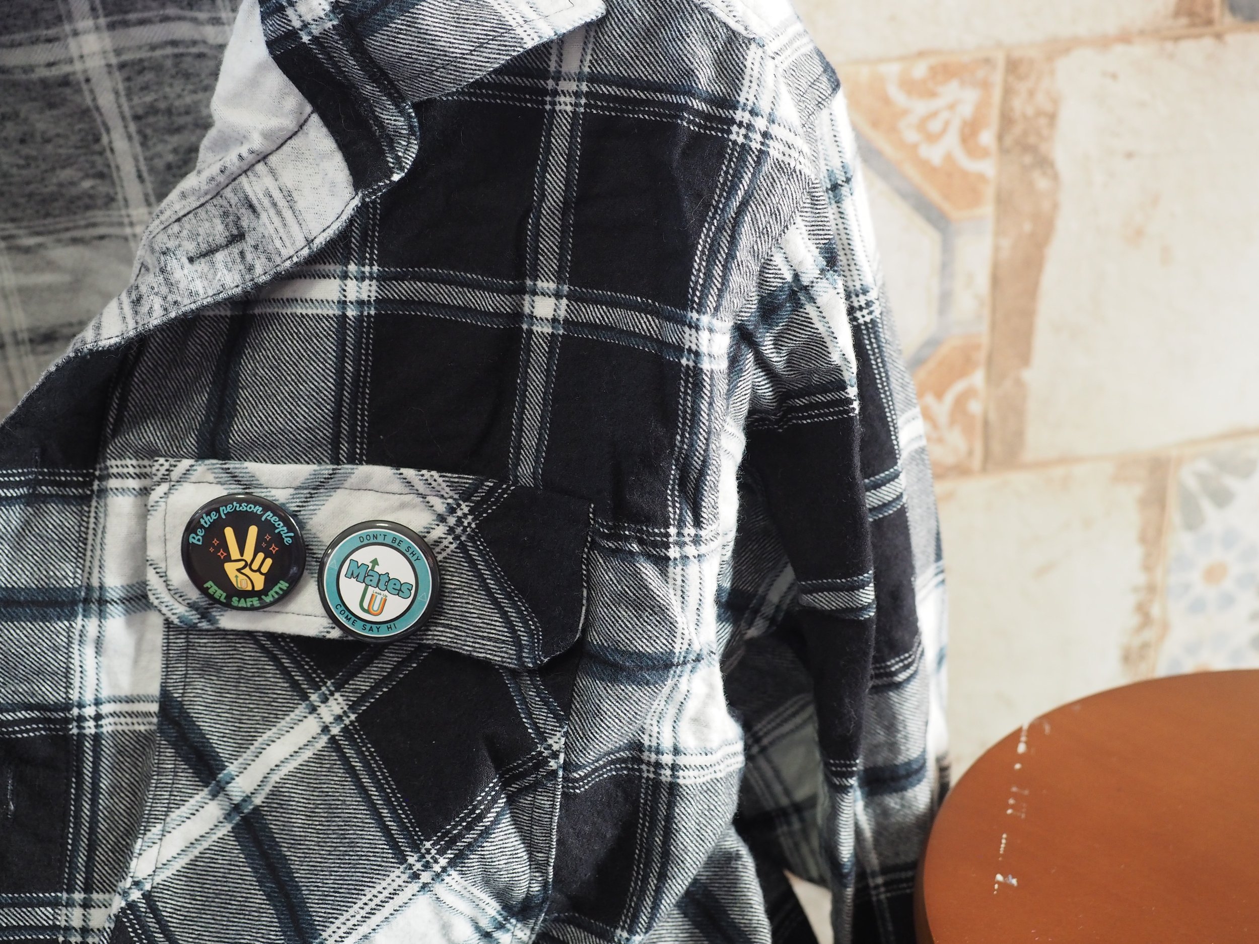 Close-up of a person wearing a black and white plaid shirt, with buttons and patches, standing near a wooden table with a textured wall in the background.