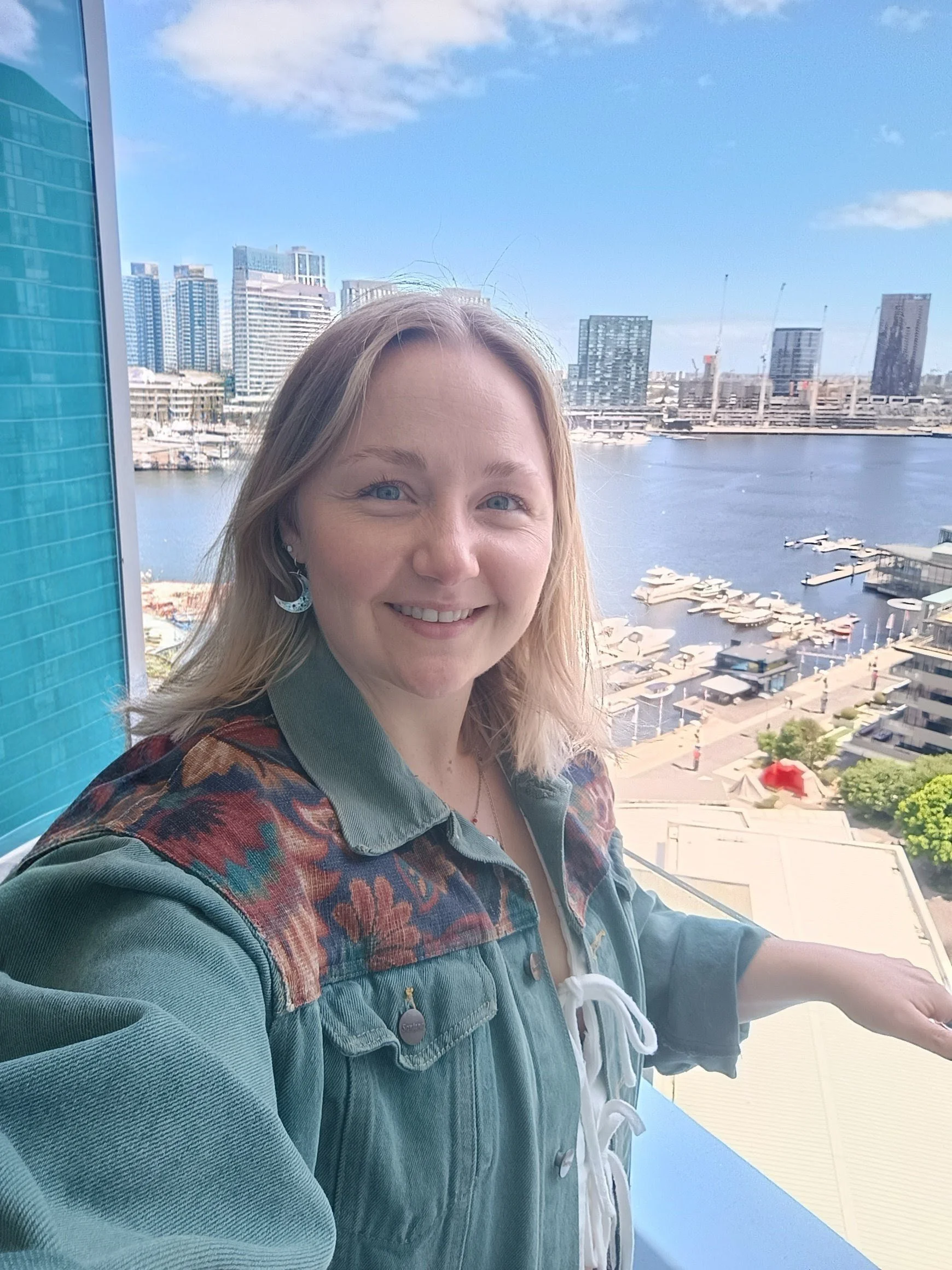 A woman smiling on a balcony overlooking a city skyline with tall buildings and a body of water.