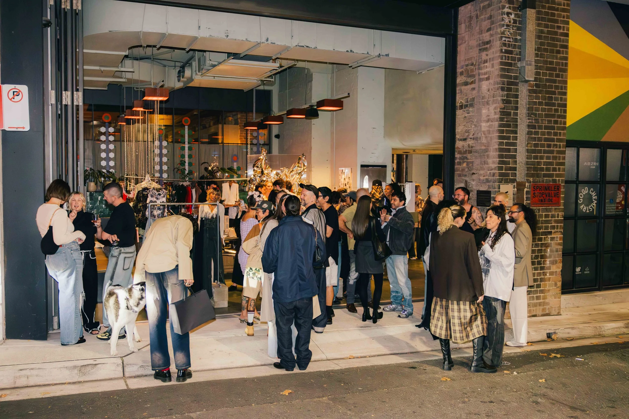 Crowd of people outside a store entrance at night, some talking and looking inside, with mannequins and clothing displays visible inside.