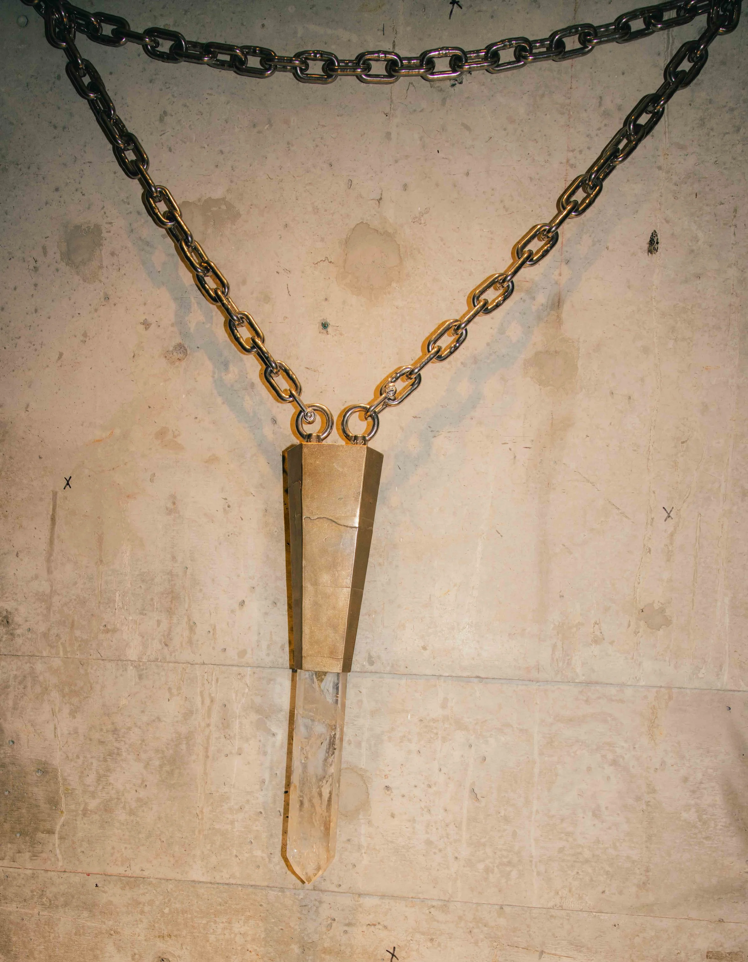 A large, metallic, crystal attached to a thick metal chain shaped like a downward-pointing arrow, hanging against a worn, beige concrete wall.