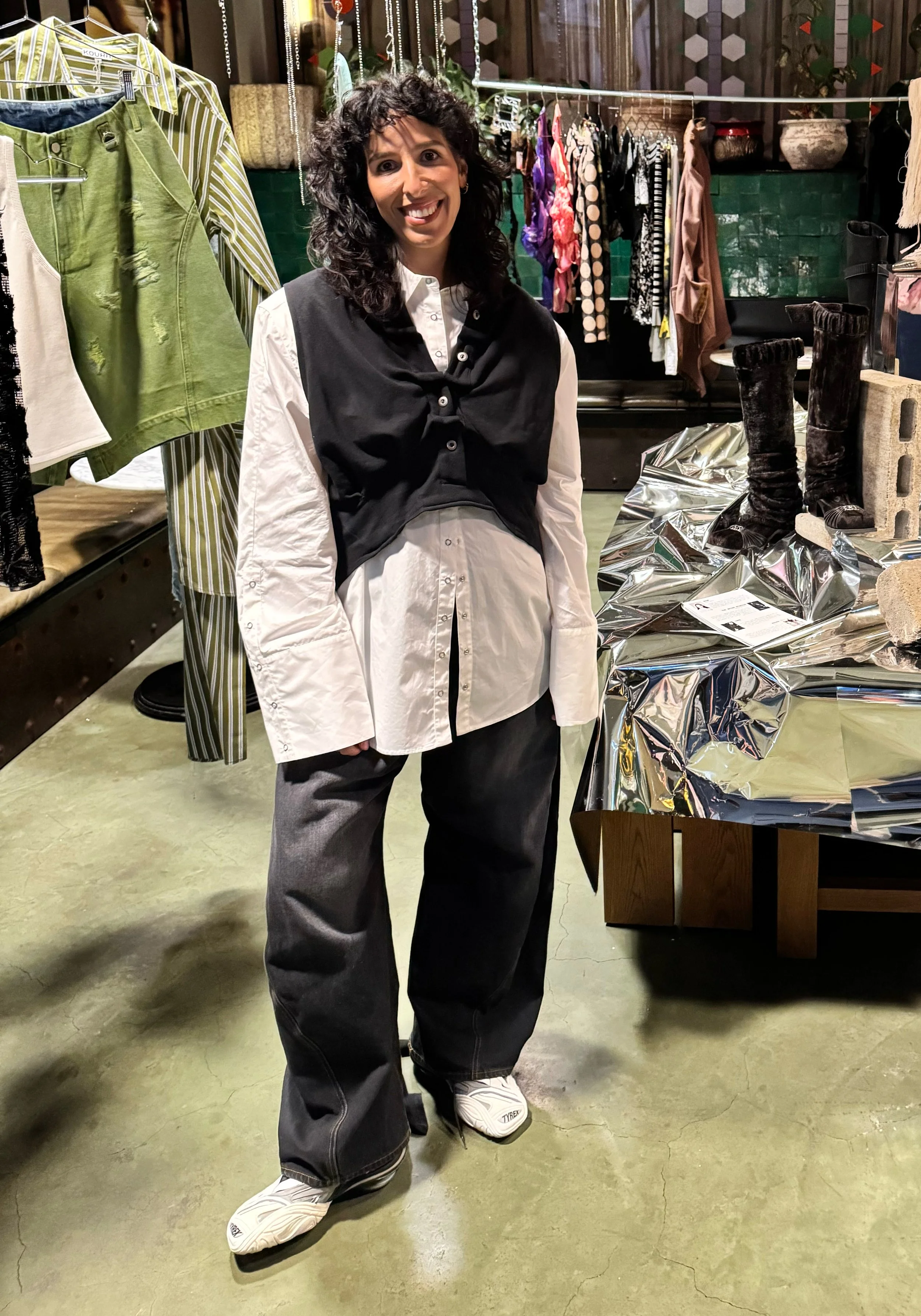 Pia Mazza, stylist, at Pinch Vol. 1, wearing a white shirt with black vest, baggy gray pants, and sneakers, surrounded by racks of clothing and accessories.