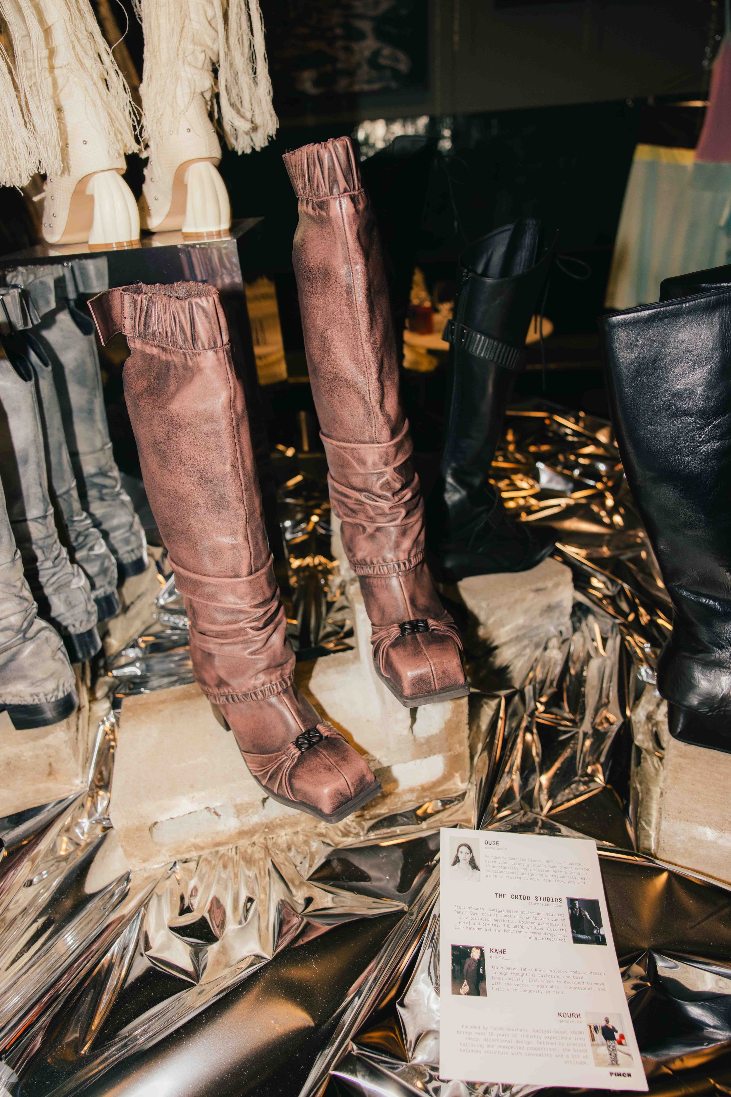 Fashion display with various boots on a glass shelf, metallic and leather finishes, with a small informational placard in the foreground.