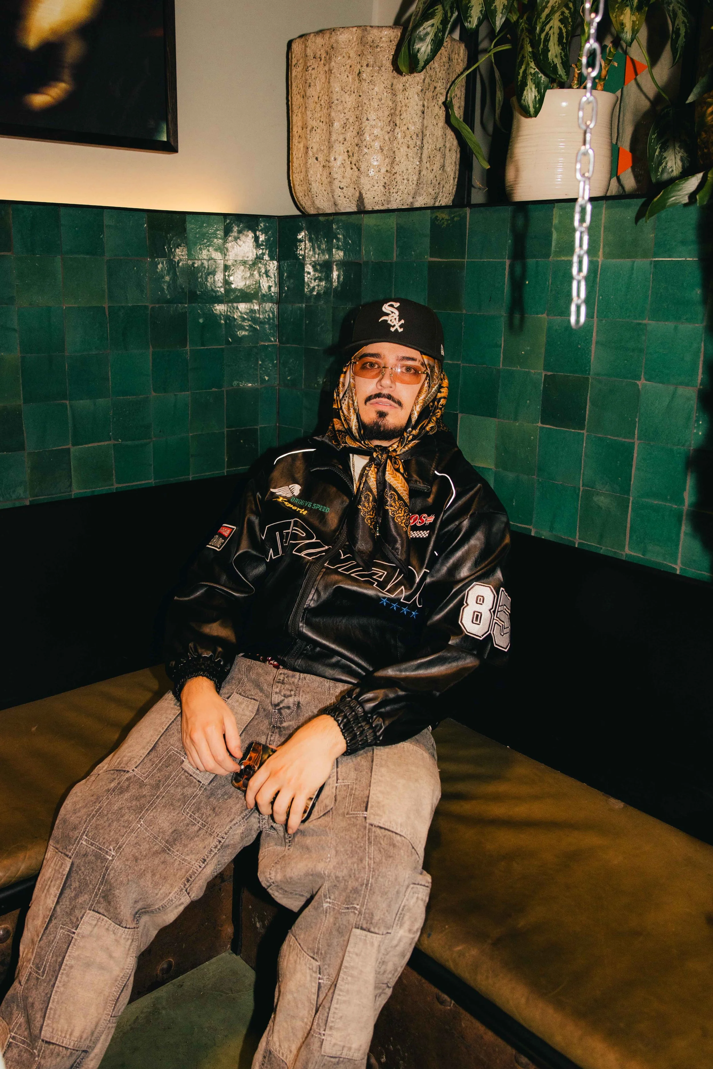 Man sitting on a banquet at Pinch Vol. 1 with green tiled wall behind him, wearing a black leather jacket, beige cargo pants, a black cap with white logo, and a patterned scarf around his neck.