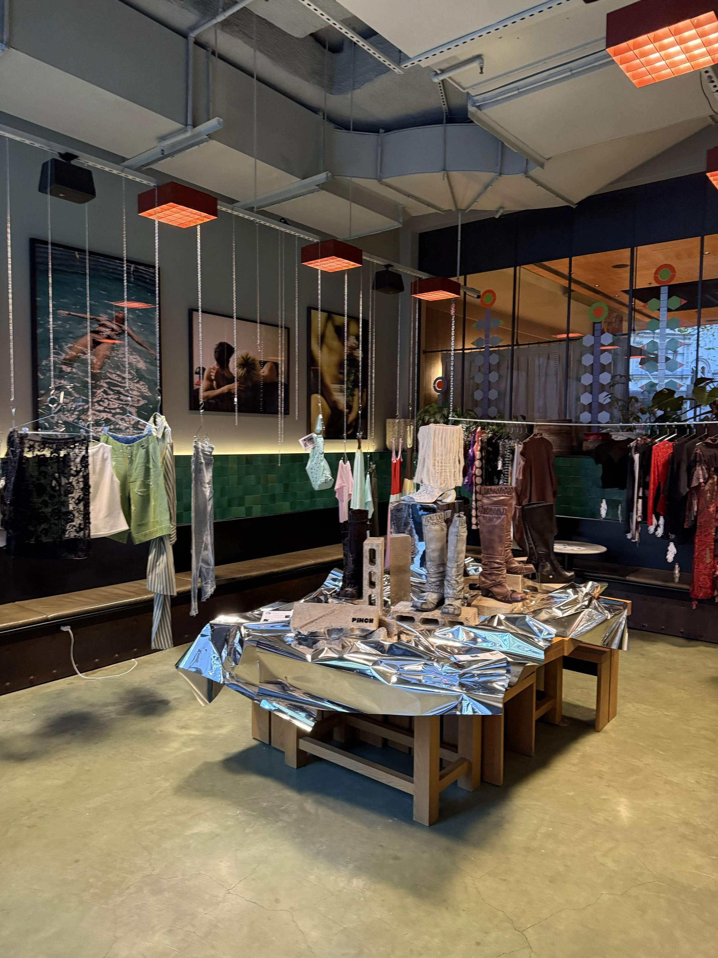 Pinch retail display with clothing items on racks and a table covered with crumpled silver foil, featuring several pairs of boots on wooden blocks at the center.