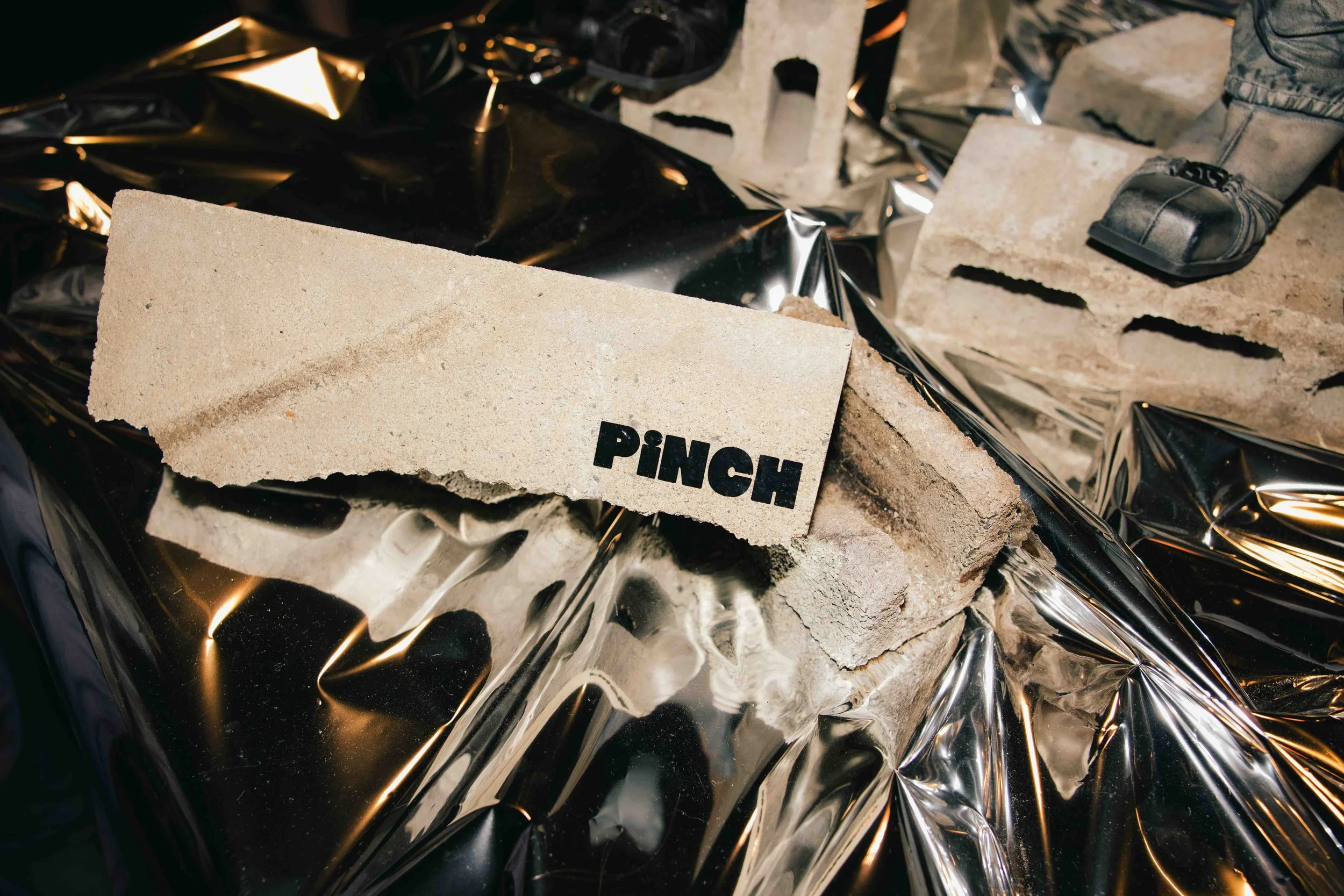 Close-up of a broken brick with the word "PINCH" on it, placed on a reflective black surface with crumpled black and shiny metallic materials underneath.