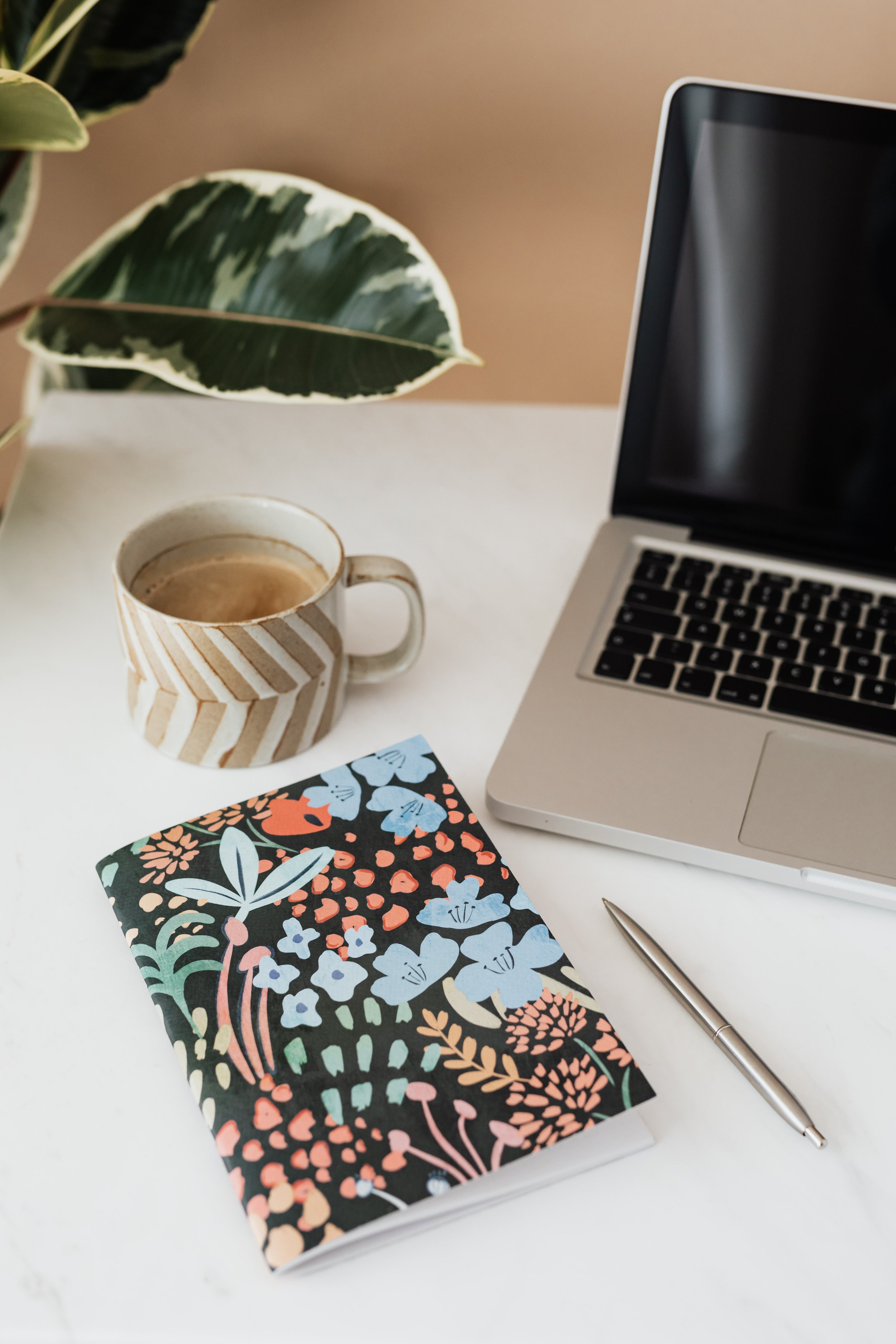 A white desk with a colorful floral notebook, a pen, a ceramic mug of coffee, a partially open laptop, and a green and white variegated plant.