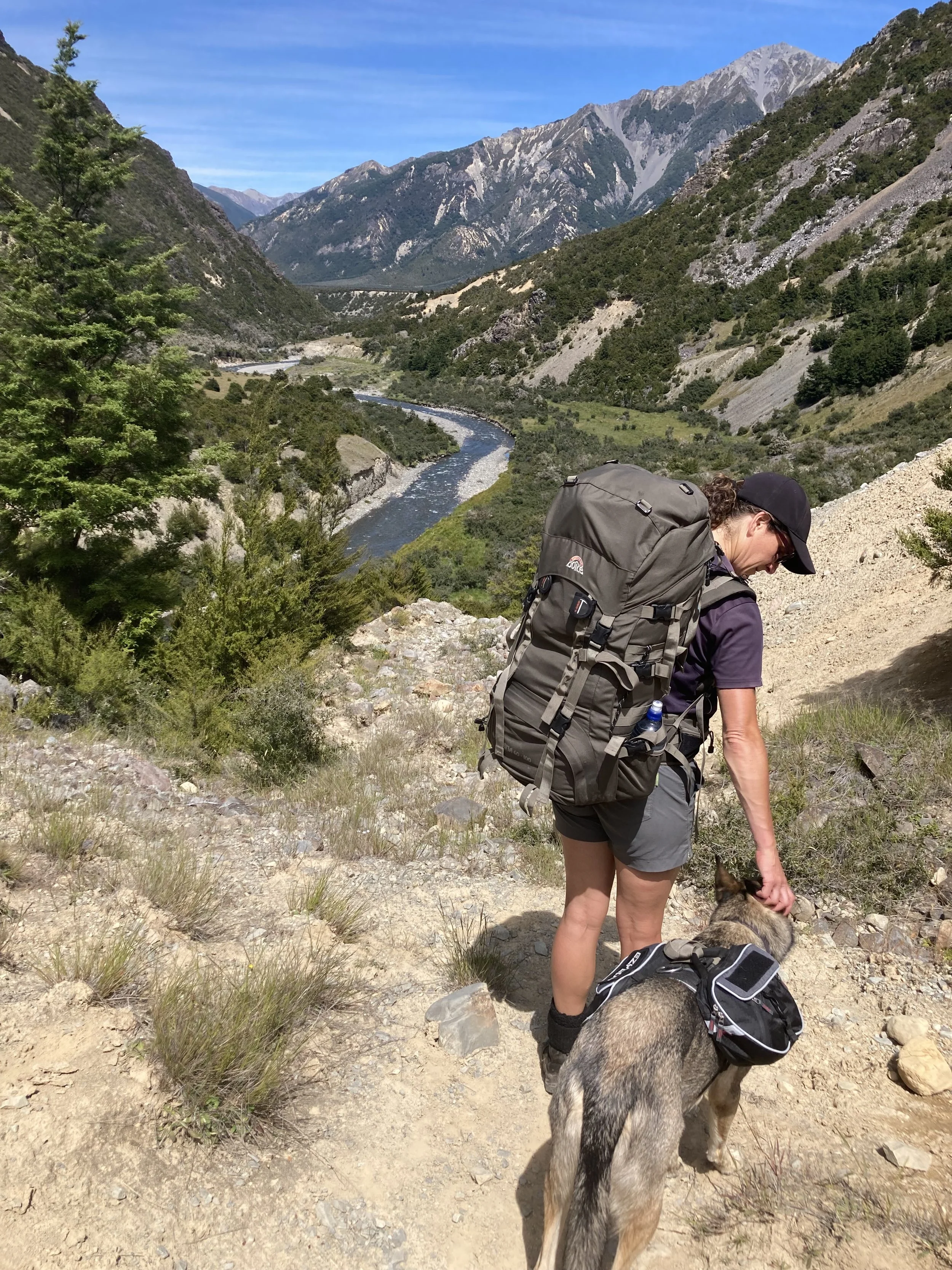 A woman wearing hiking gear and a cap stands on a rocky trail in a mountainous area, petting a dog with a scenic river and mountain landscape in the background.