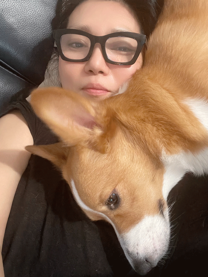 A woman with glasses lying down next to a brown and white dog, both appearing relaxed and close together.