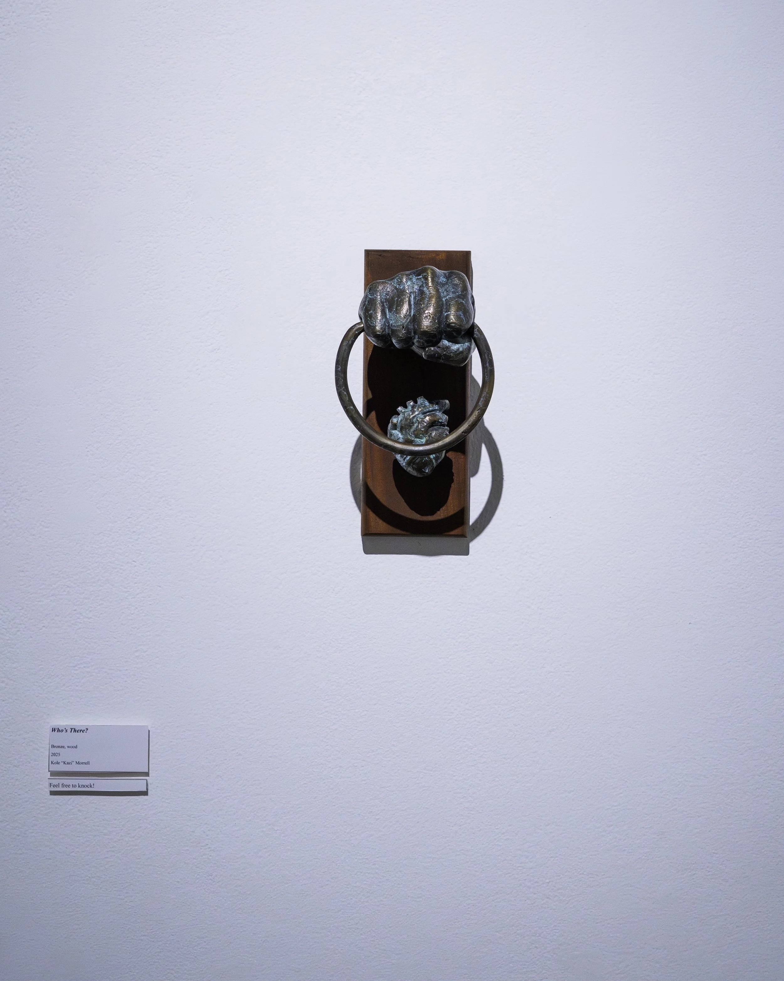 Bronze sculpture of a fist gripping a ring mounted on a wooden base displayed against a white wall.