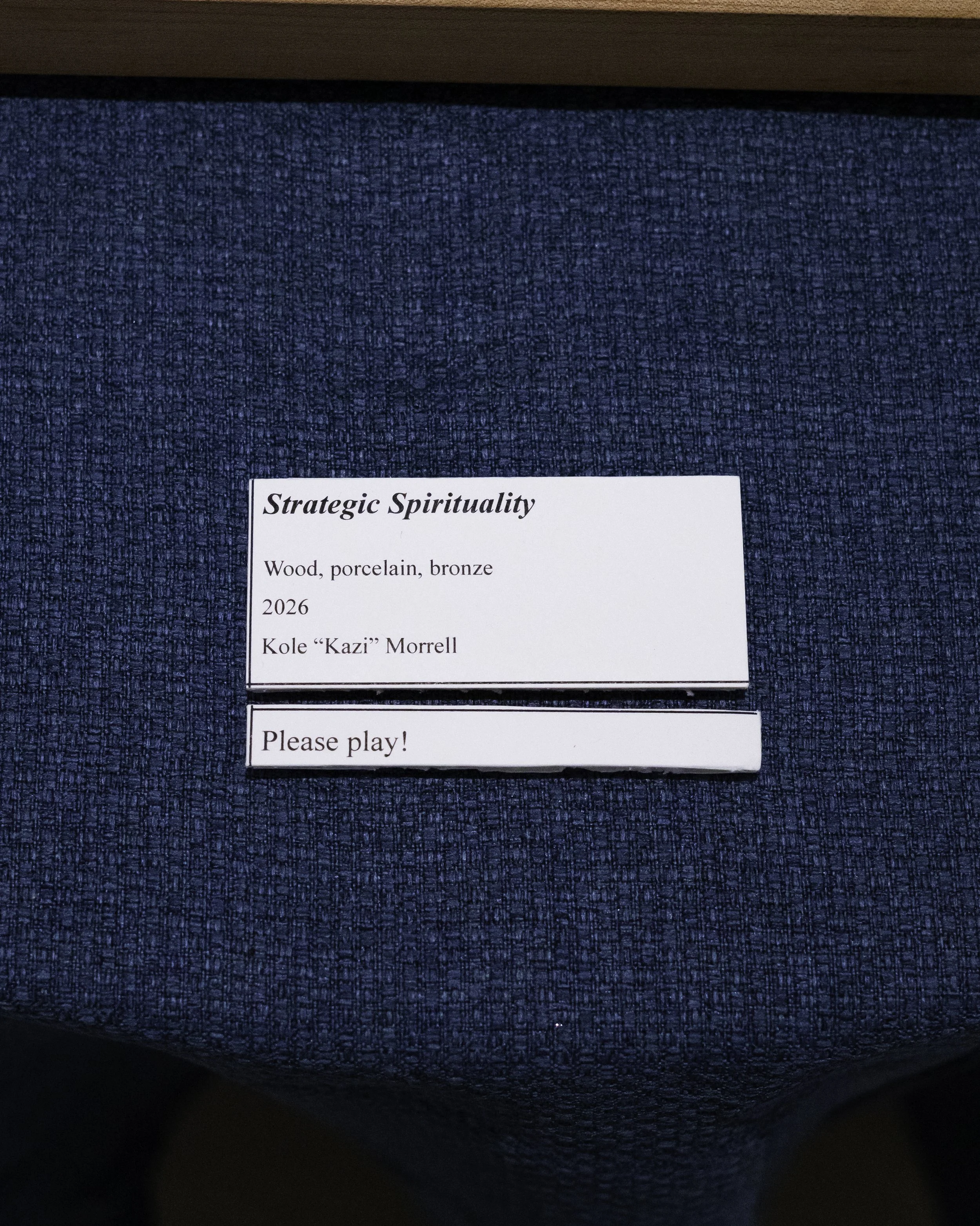 Close-up of a display card labeled 'Strategic Spirituality,' describing a piece made of wood, porcelain, and bronze by Kole 'Kazi' Morrell from 2026, placed on a blue textured surface with a wooden edge.