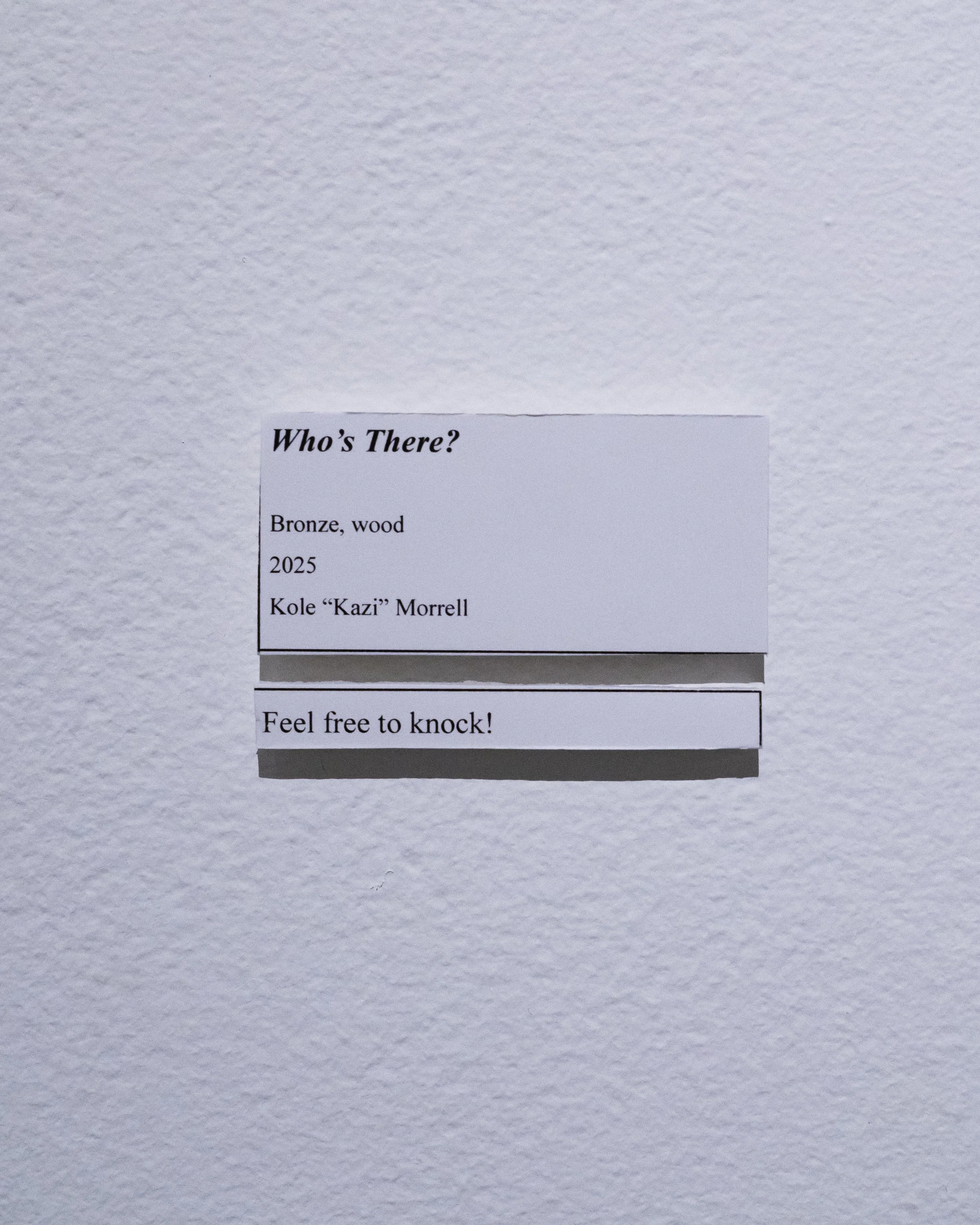 A white wall with a small, partially visible poster or paper with text that reads 'Who's There?' and information about bronze wood by Kole 'Kazi' Morrell, dated 2025, and a 'Feel free to knock!' message.
