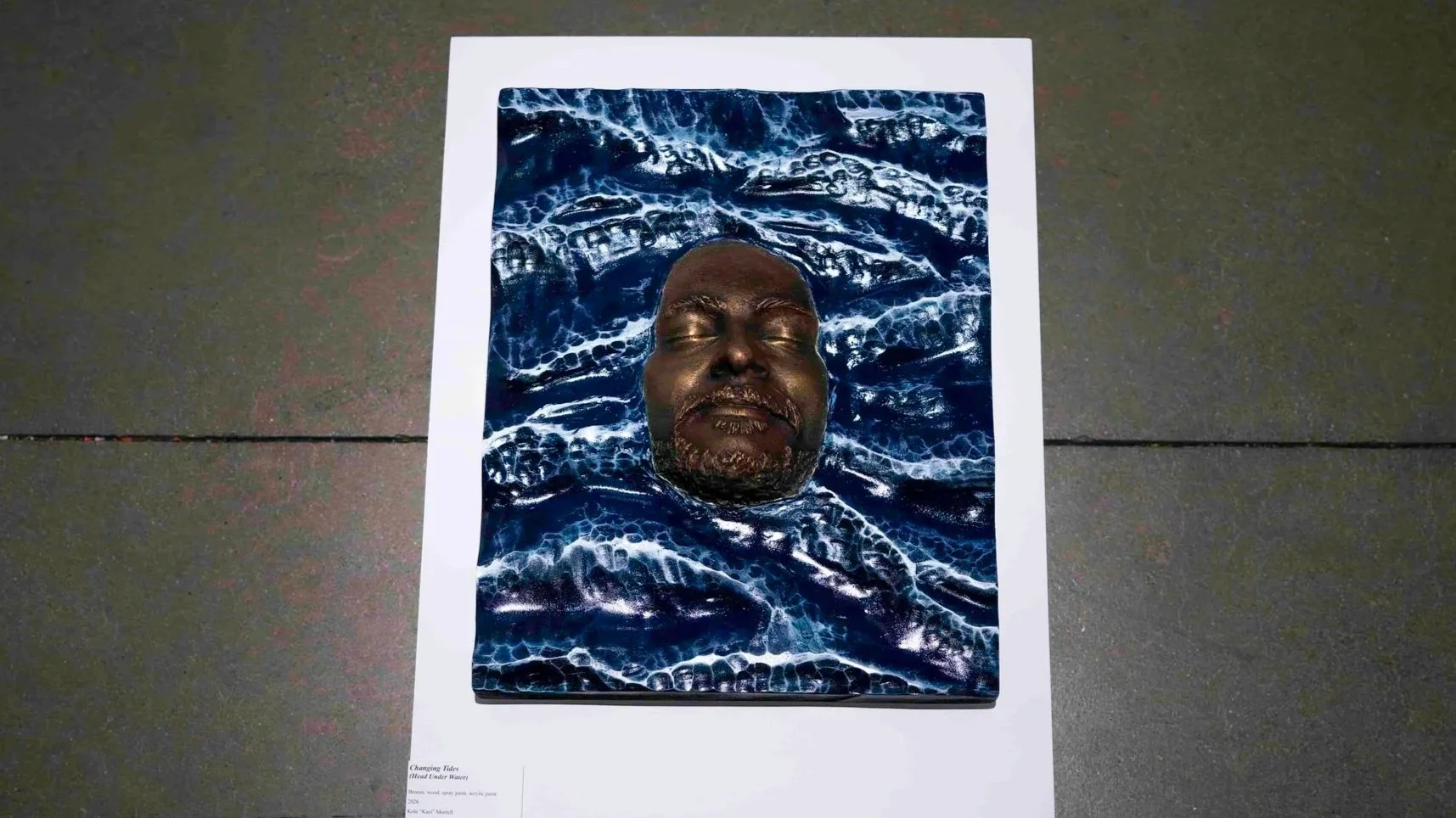 Art piece featuring a man's face submerged in water, with his eyes closed, surrounded by dark blue waves and foam.