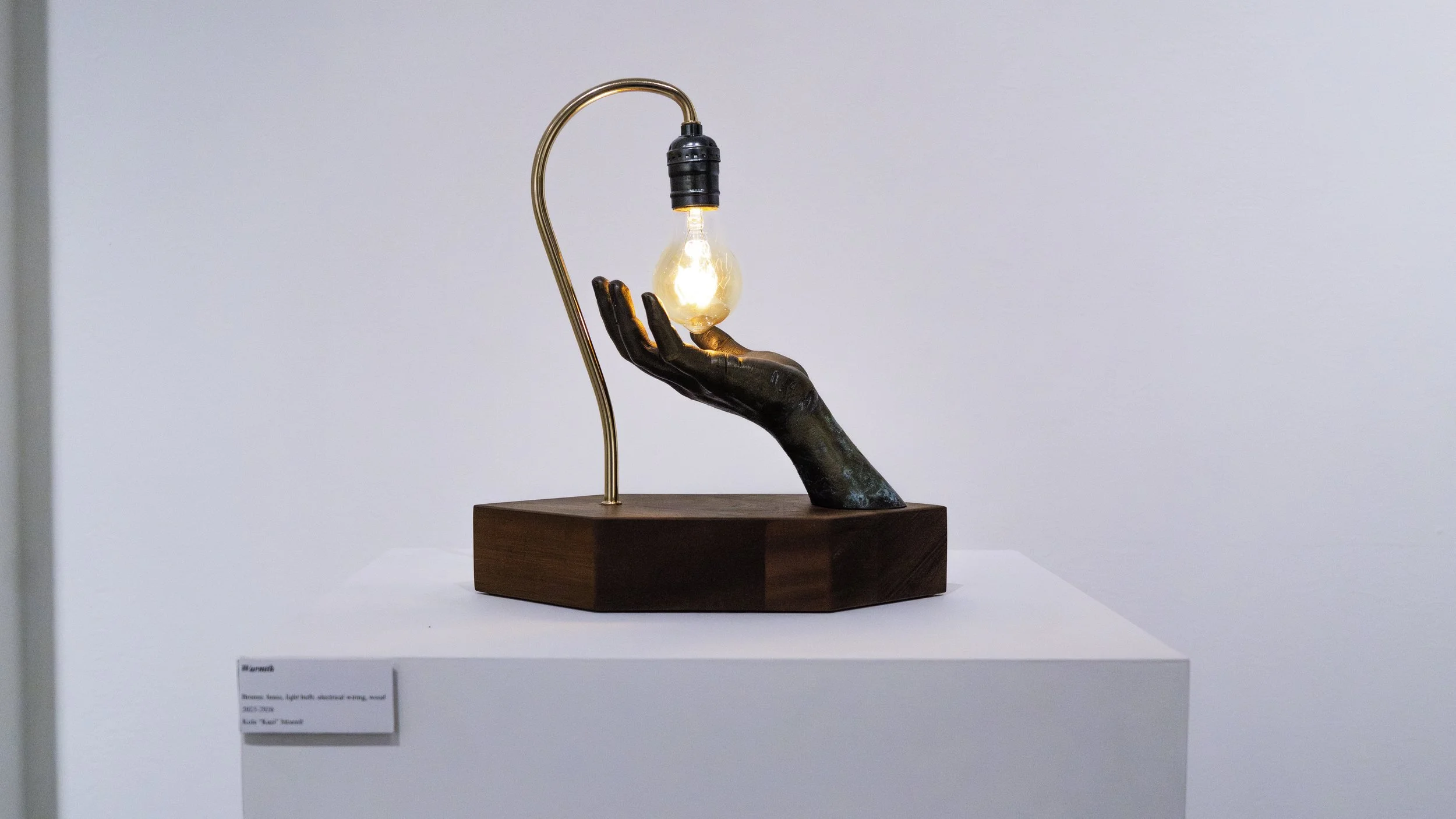A sculpture of a hand with a dark patina, supporting a glowing light bulb, with a copper-colored flexible lamp arm extending from behind. The sculpture is mounted on a wooden base, displayed in a gallery setting.