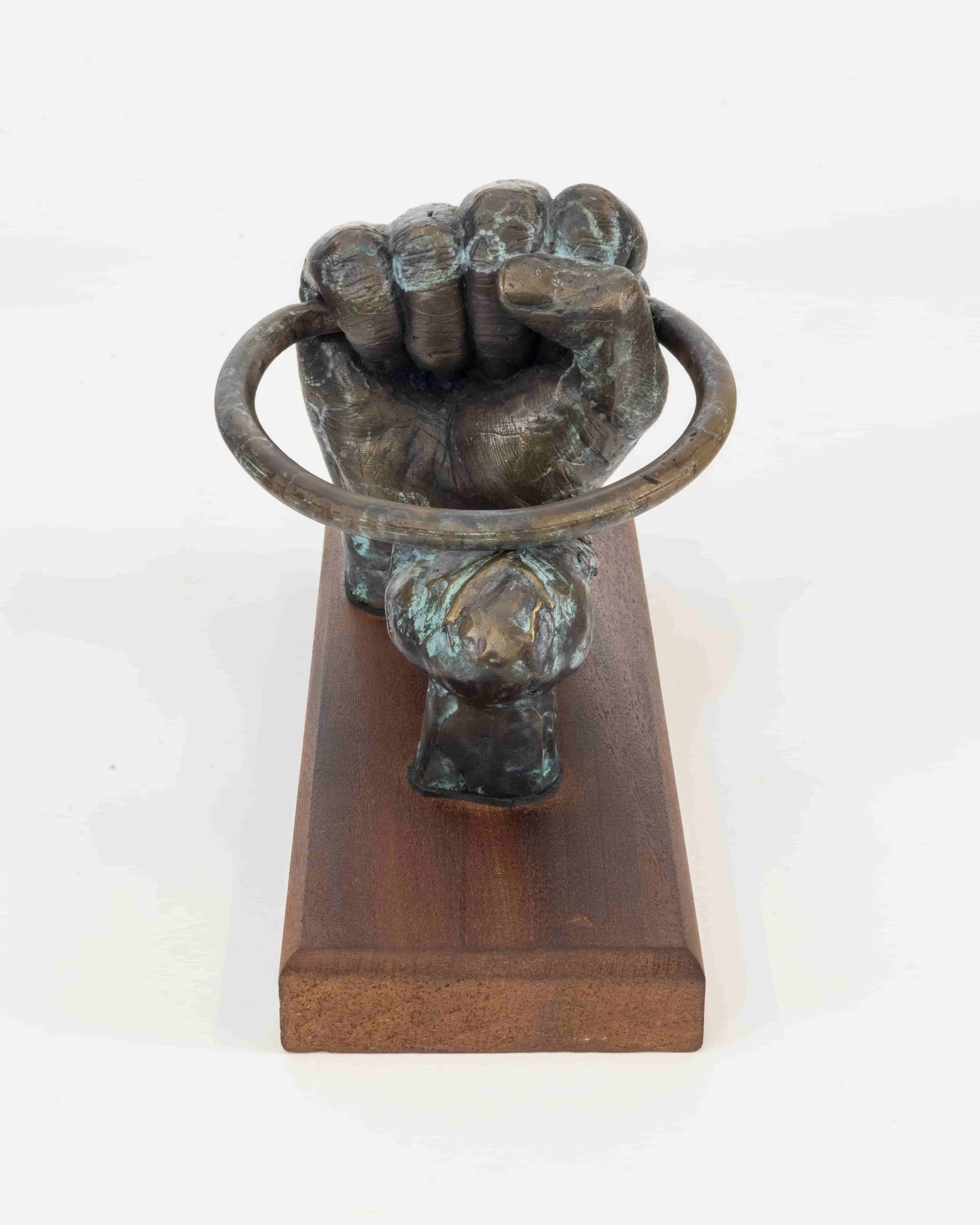 Bronze sculpture of a person's clenched fist gripping a ring, mounted on a wooden base.