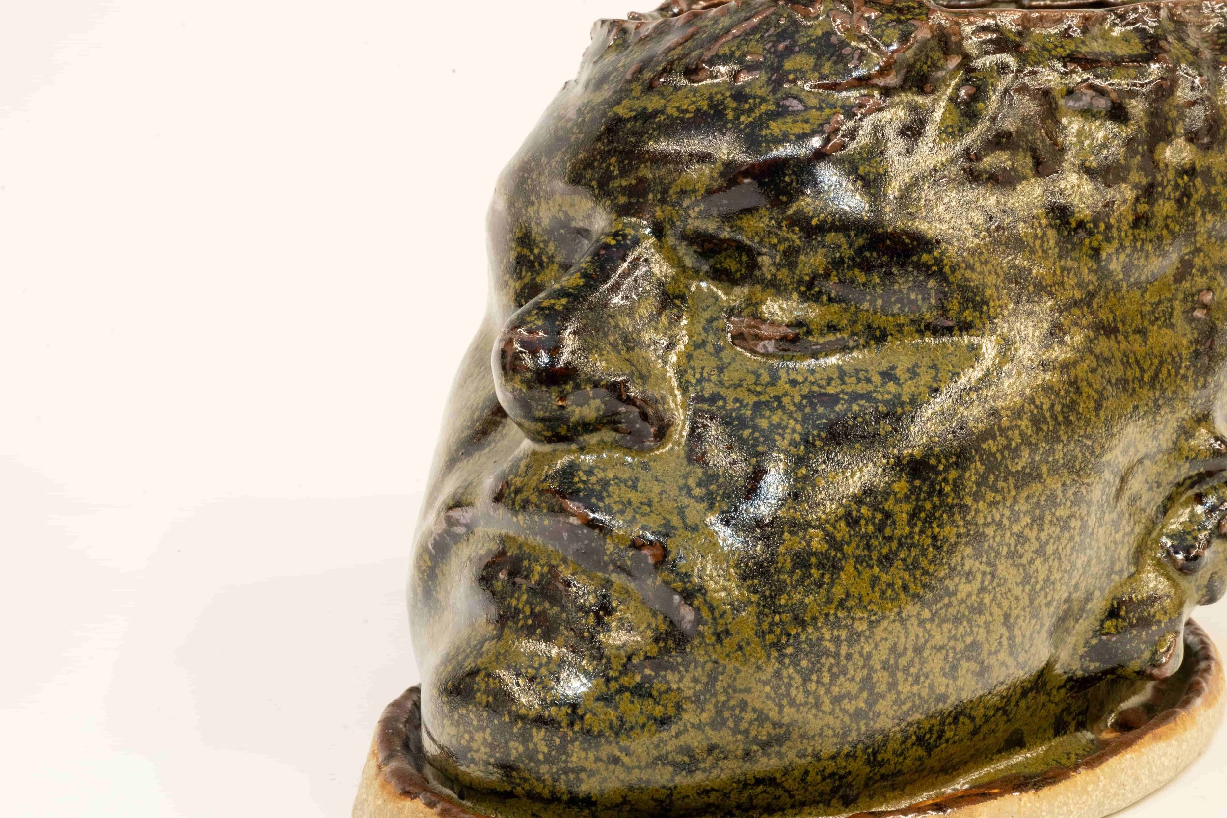 Close-up of a greenish-brown sculpture of a person's face, showing detailed features such as closed eyes, nose, and lips, with a textured surface.