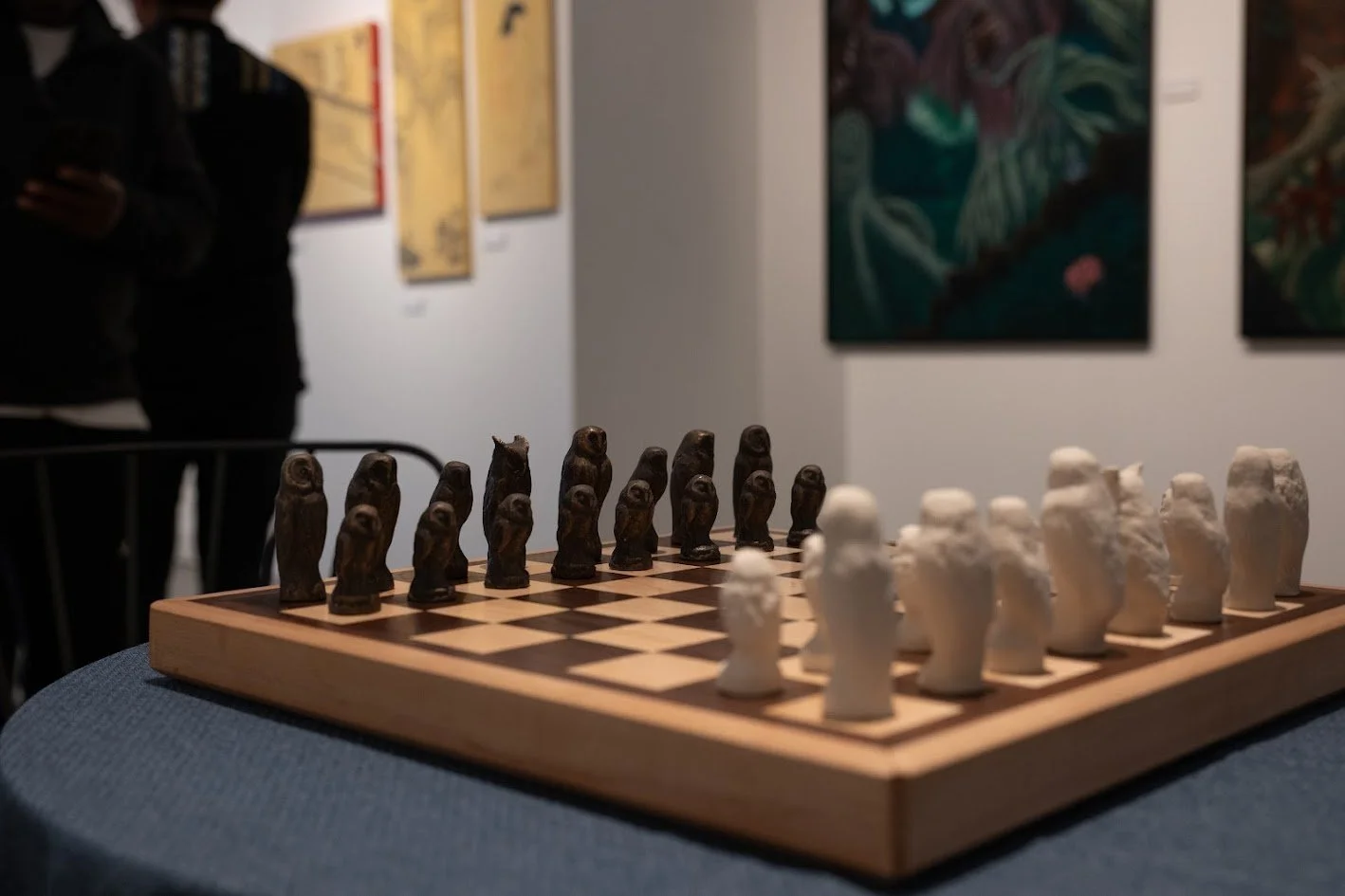 A chessboard with black and white chess pieces displayed in an art gallery with paintings on the wall in the background.
