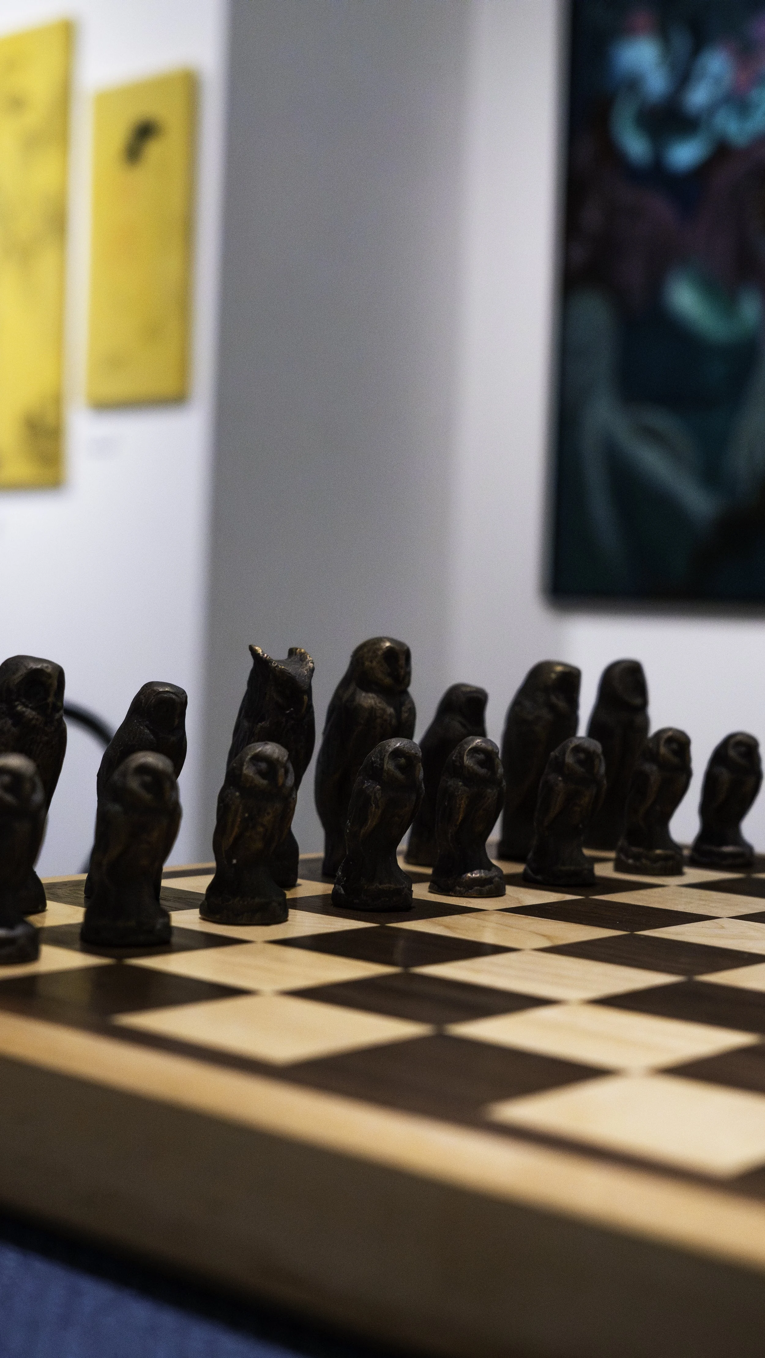 Wooden chess pieces arranged on a chessboard, with artwork visible on the wall behind.