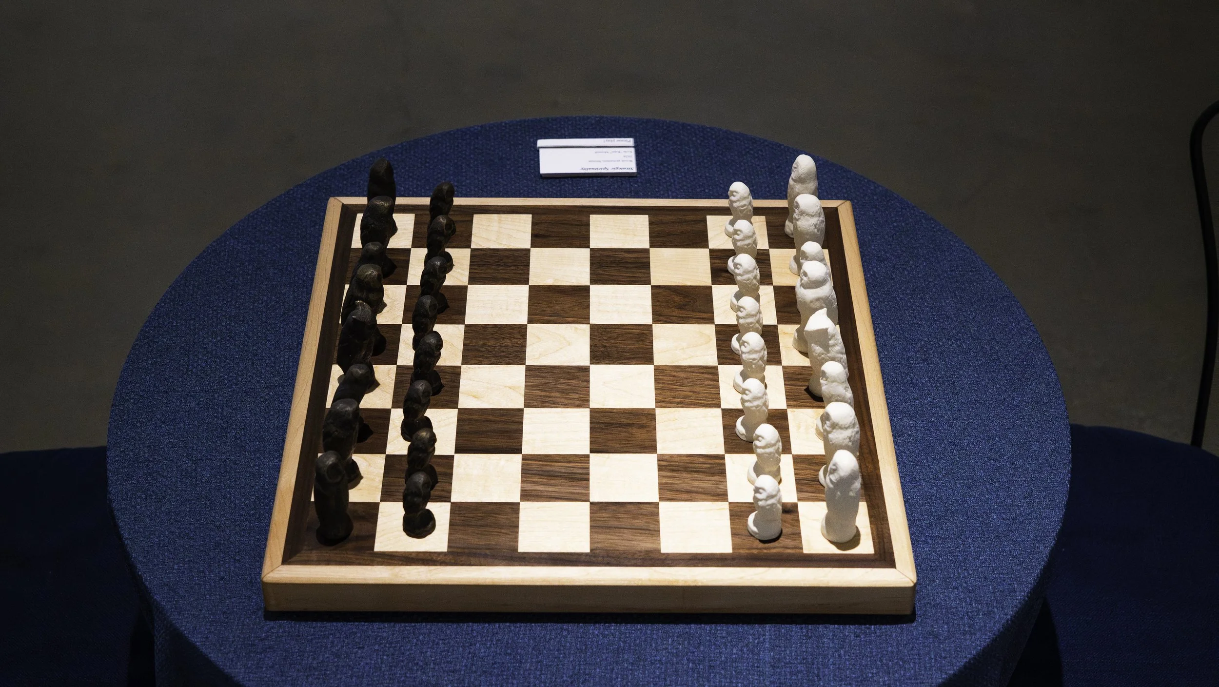 A chessboard set up for a game with black and white pieces in their starting positions on a blue round table with a small card in the background.