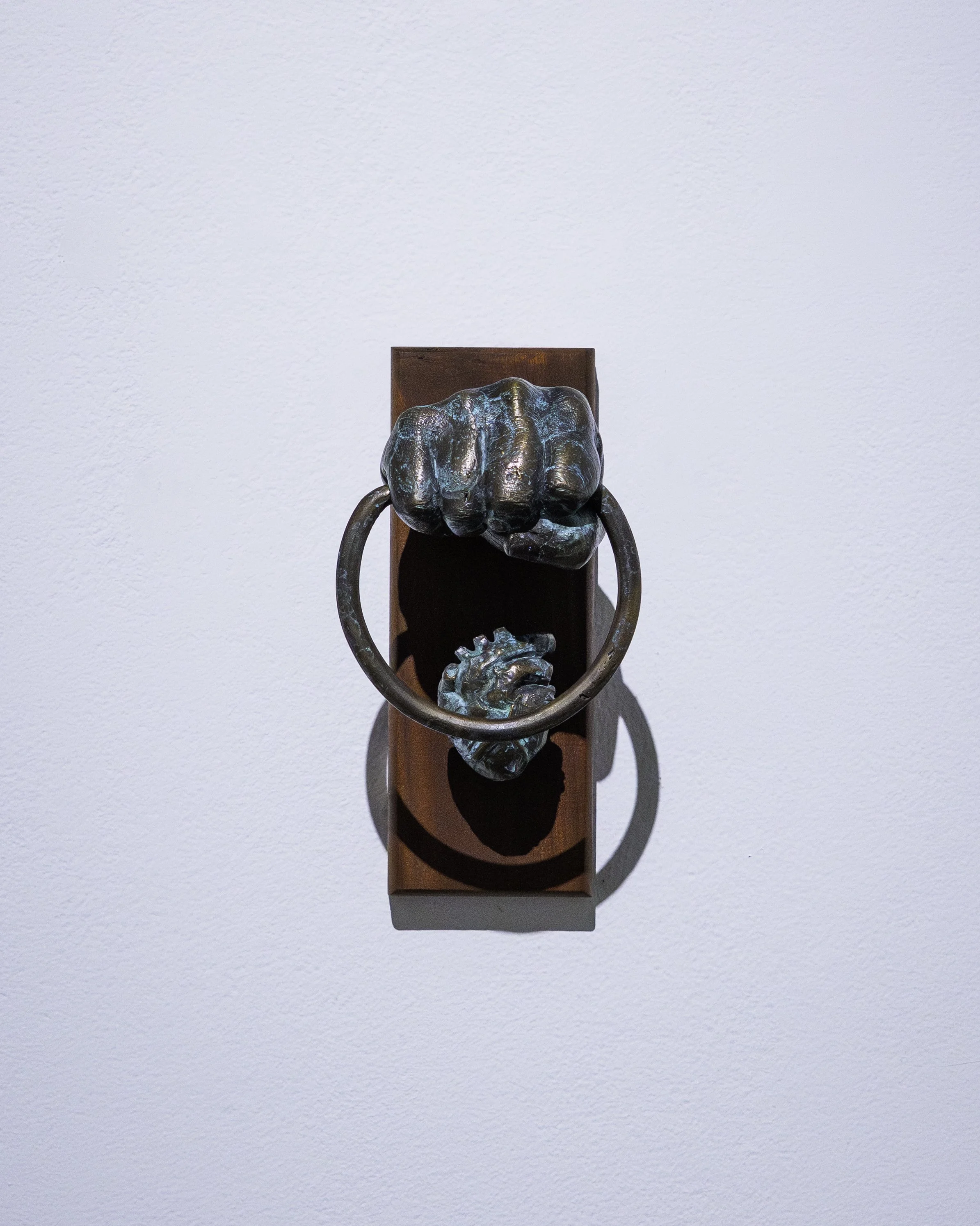 A bronze hand sculpture gripping two large metal rings mounted on a wooden plaque against a white wall.