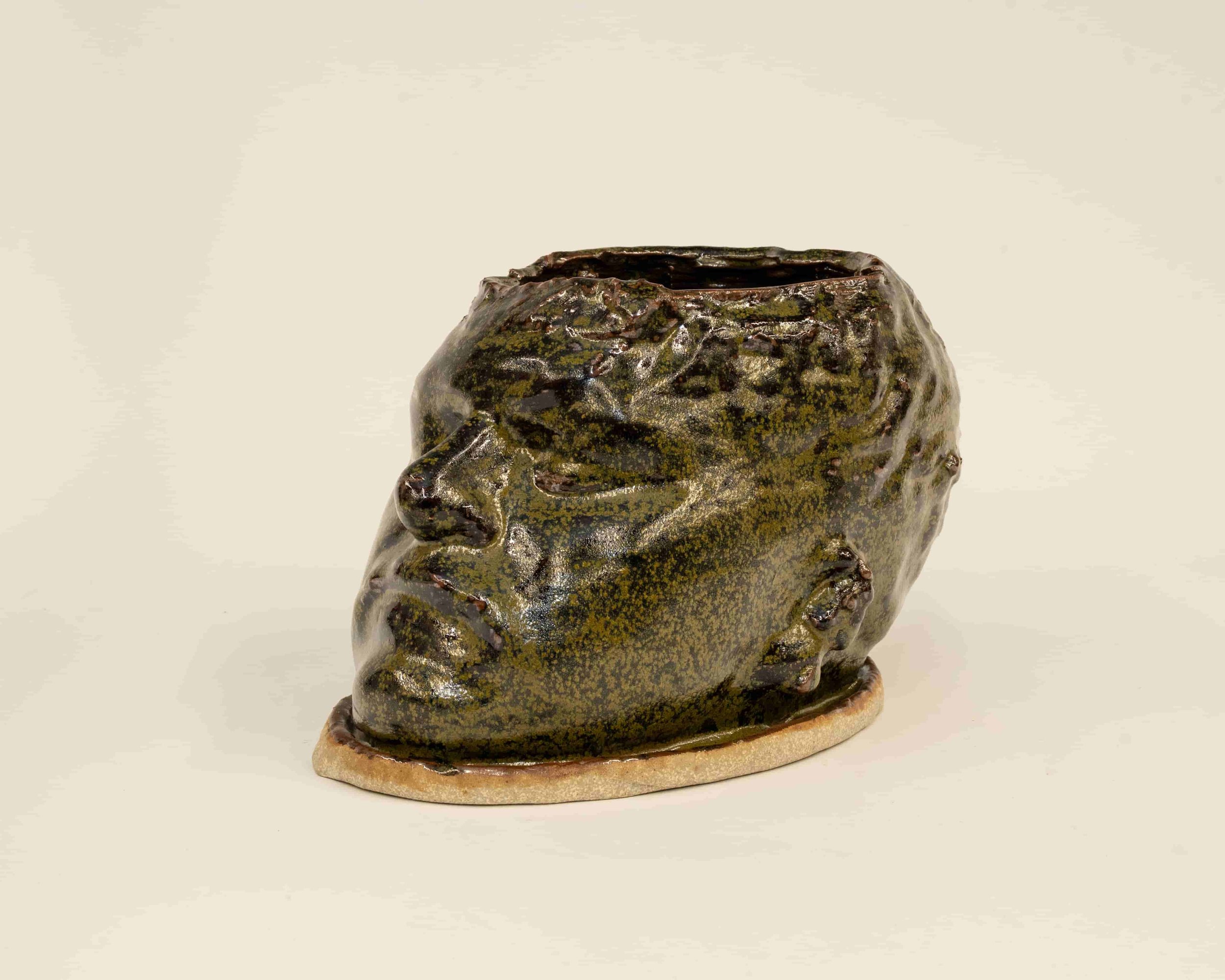 A ceramic sculpture resembling a face with closed eyes and textured hair, painted in dark green, brown, and black, mounted on a rectangular base.