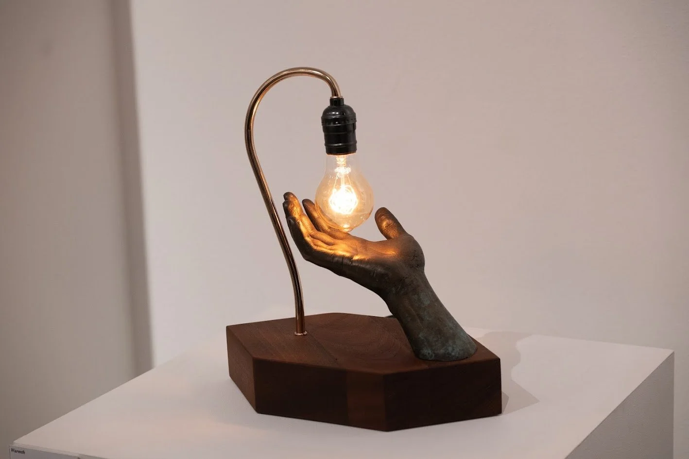 A unique lamp featuring a hand sculpture with a dark patina holding a glowing lightbulb, mounted on a wooden base.