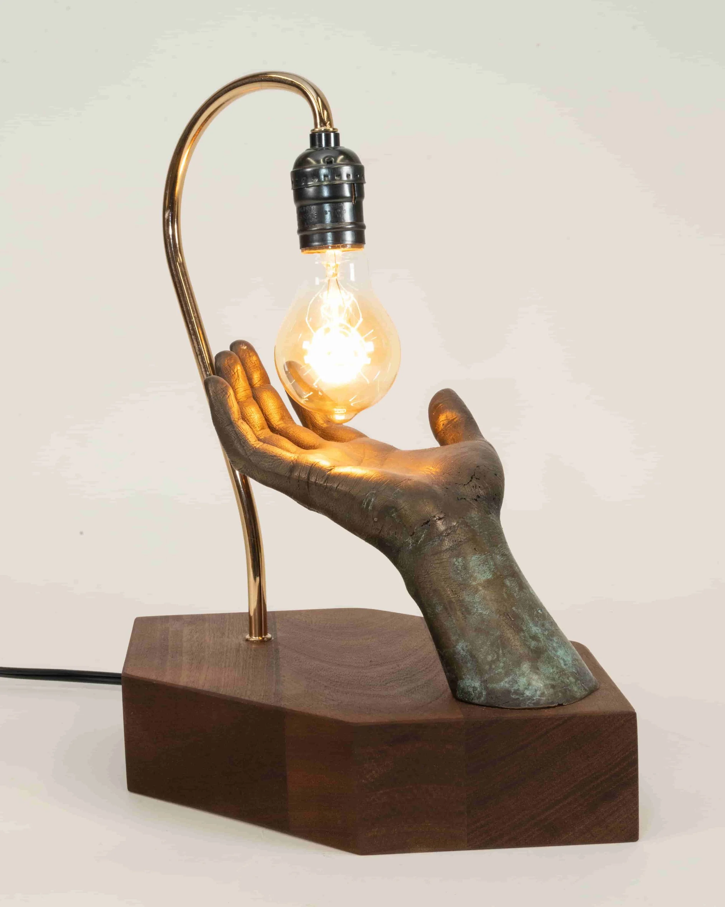 A sculpture of a hand holding a lightbulb creates a lamp, mounted on a wooden base.