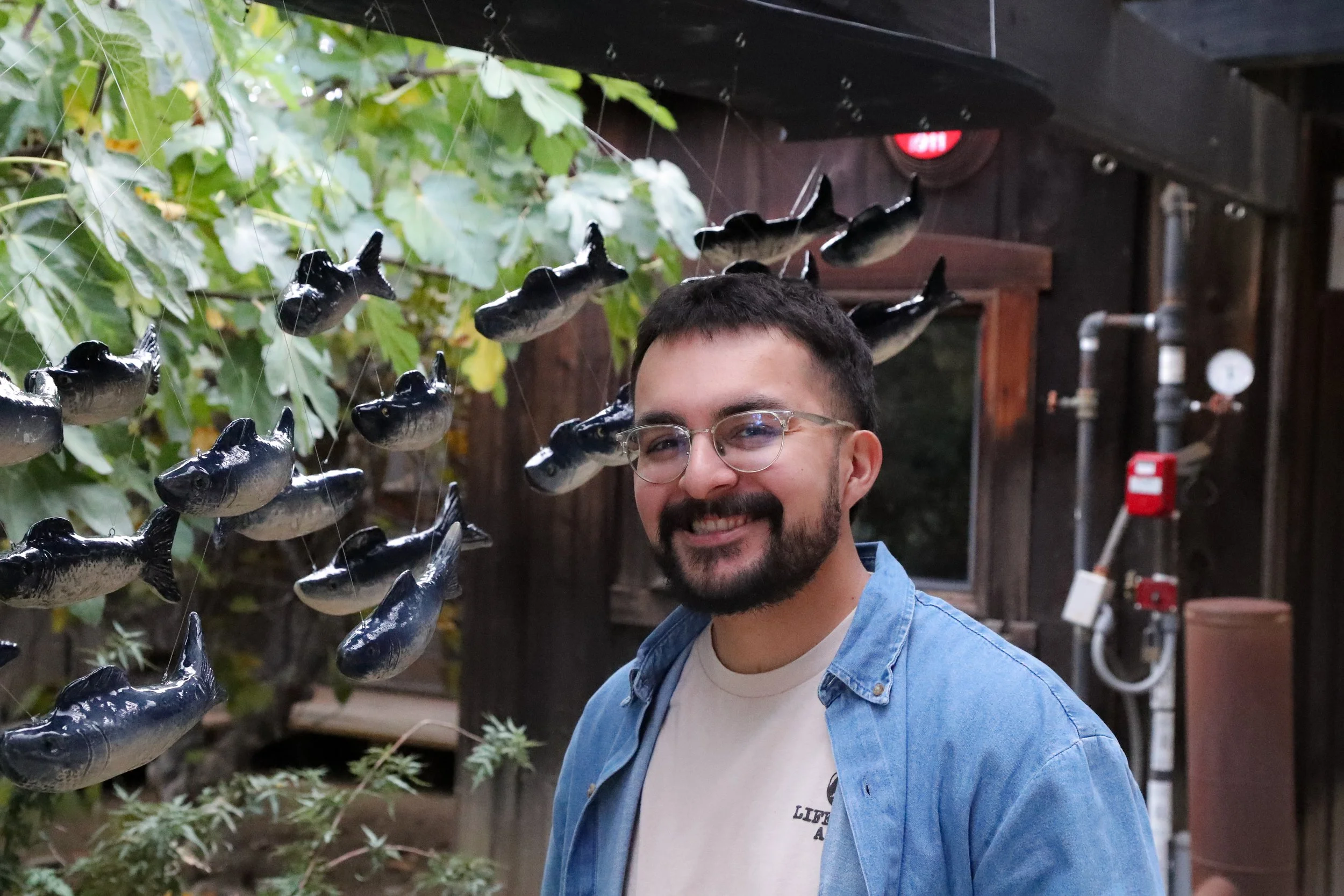 A smiling man with glasses and a beard posing in front of a display of black fish sculptures hanging from above, with greenery and wooden structures in the background.