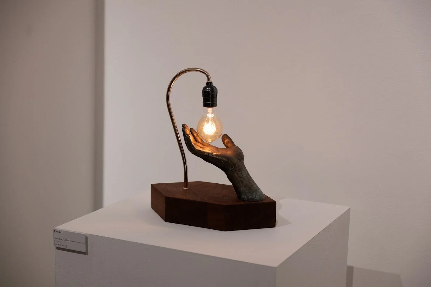 A sculpture of a black hand holding a glowing light bulb, with a curved metallic arm supporting the bulb, mounted on a wooden base, displayed in an art gallery.