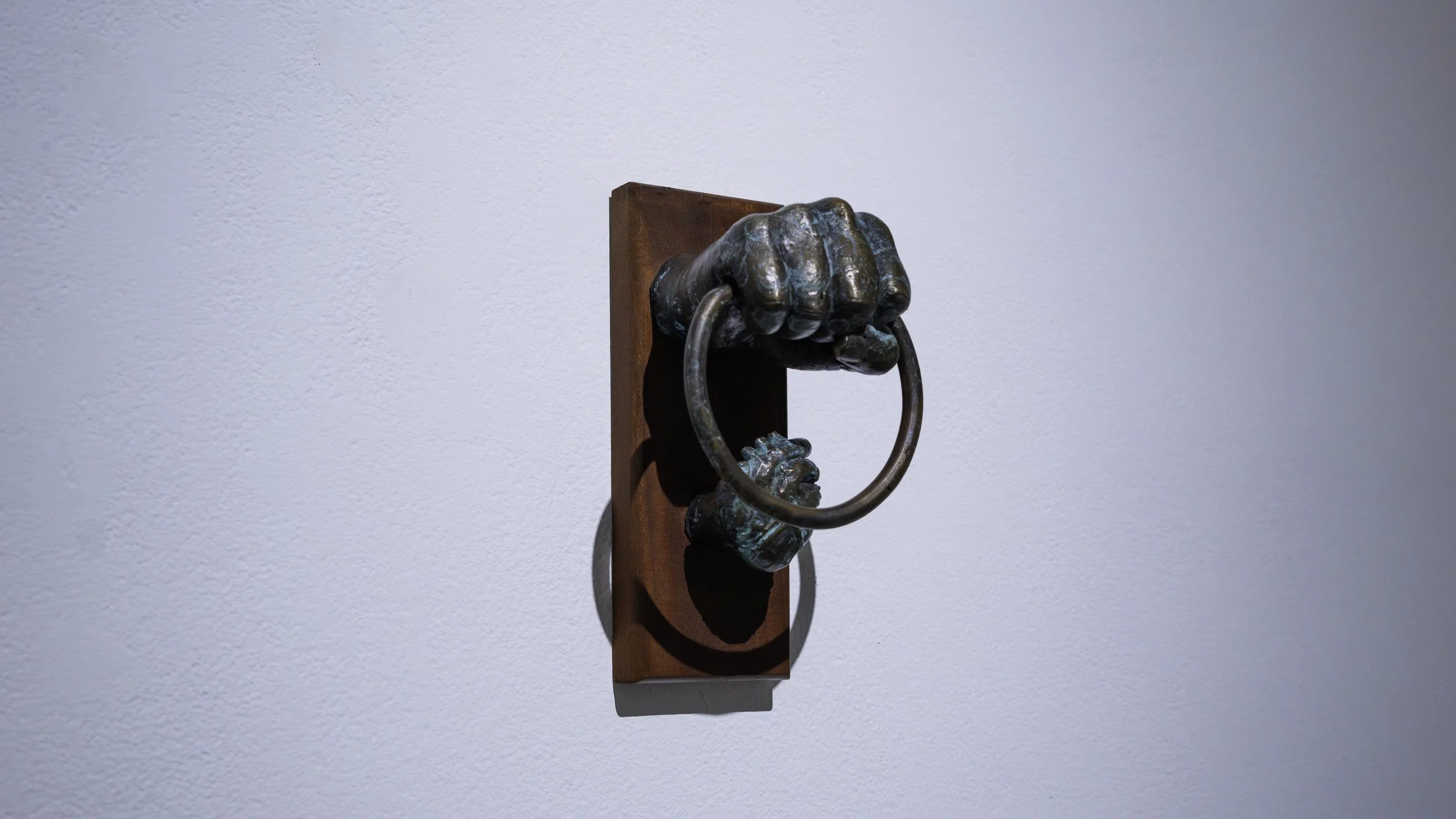 Bronze sculpture of a hand gripping a ring mounted on a wooden plaque, positioned on a white wall.