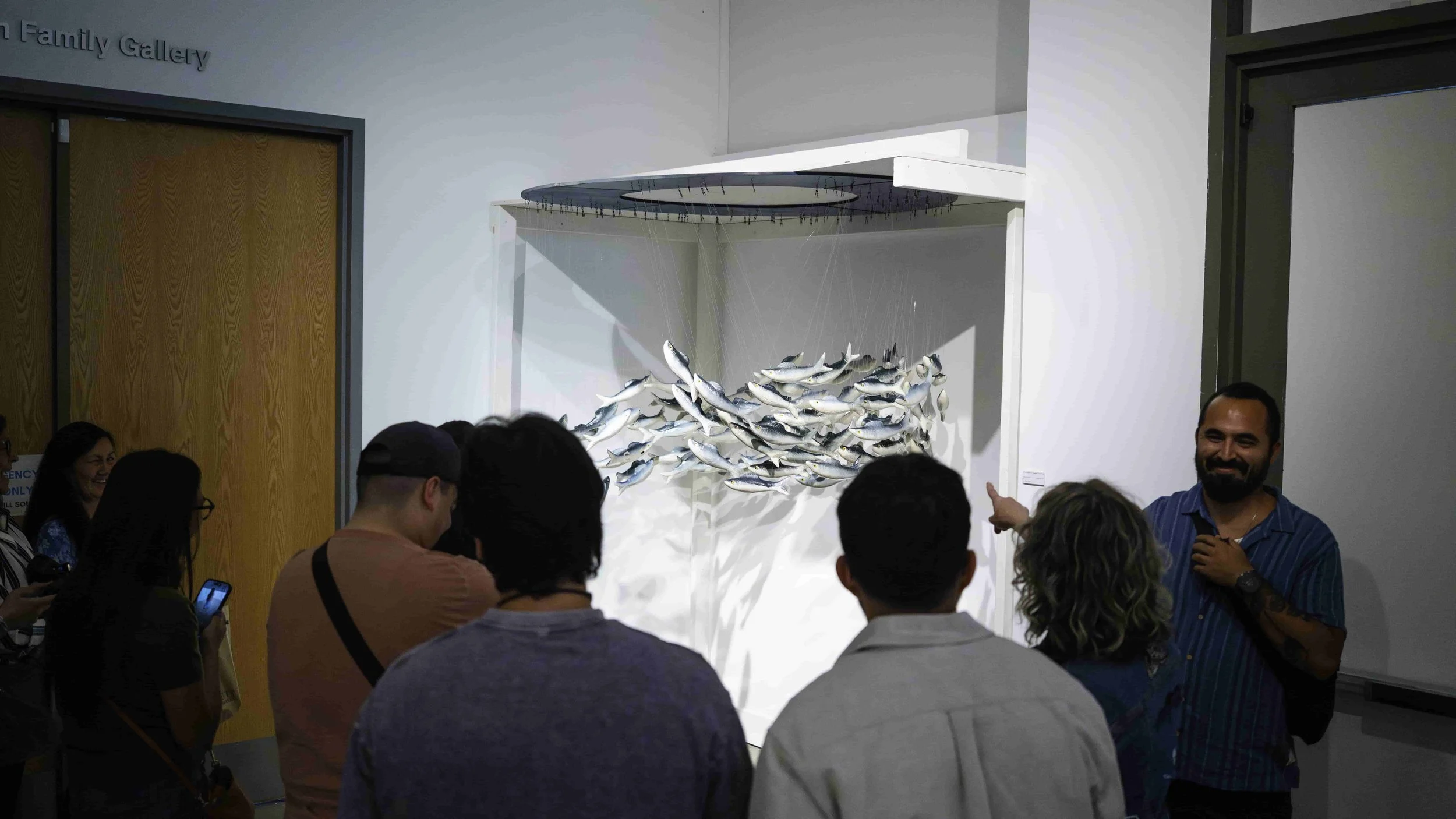 Group of people gathered in front of an art installation of a school of fish hanging from the ceiling at a museum or gallery.