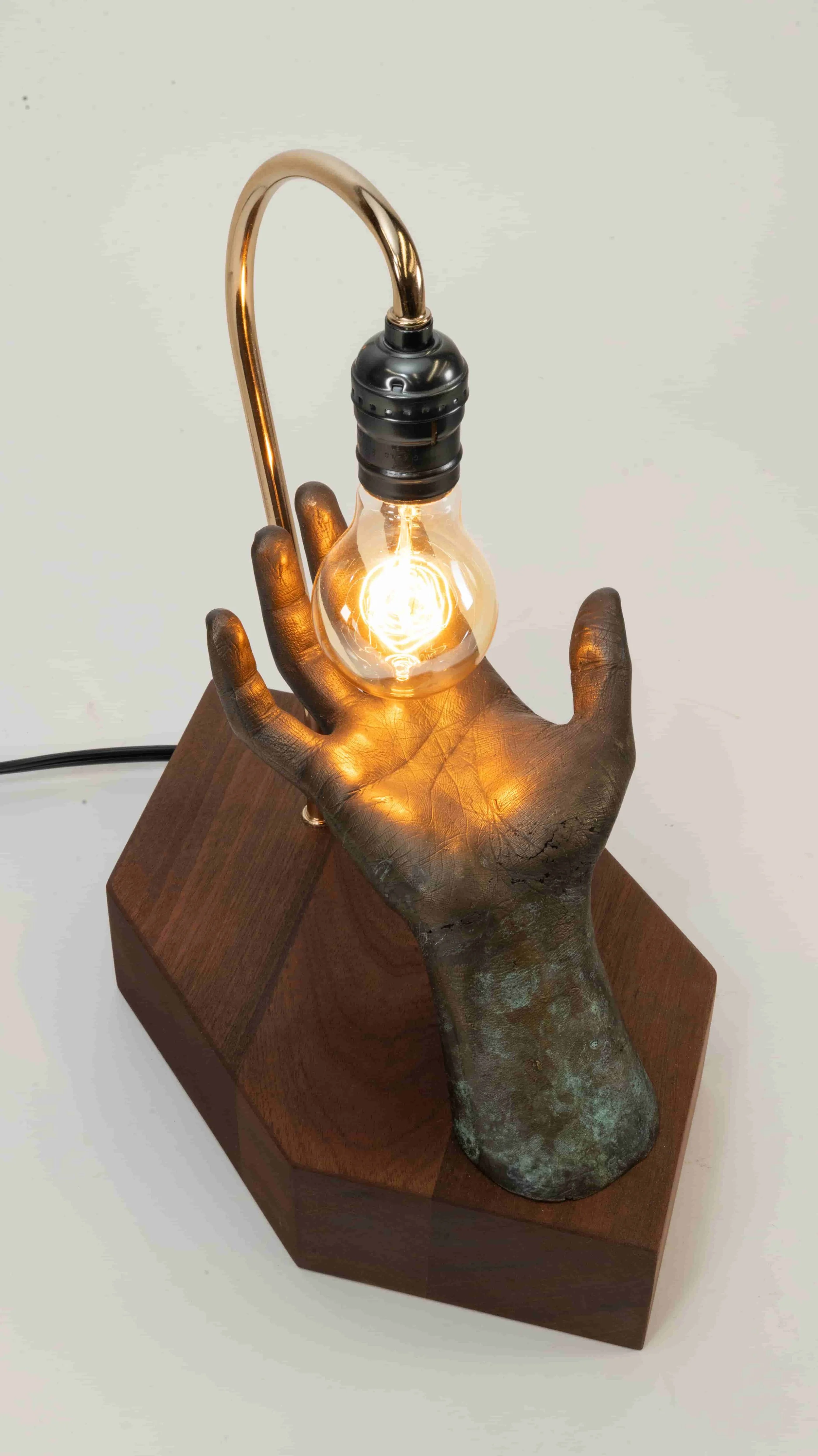 Sculpture of a metallic hand holding a light bulb with a filament glowing, mounted on a wooden base.