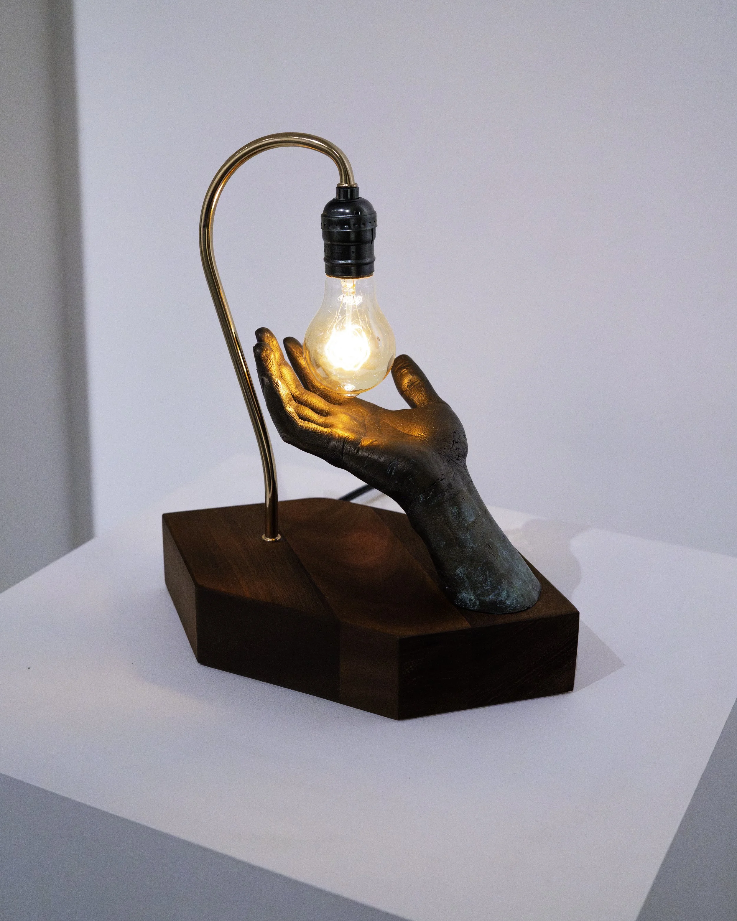 A sculpture of a human hand holding a light bulb, with a metal stand and wooden base, against a plain background.