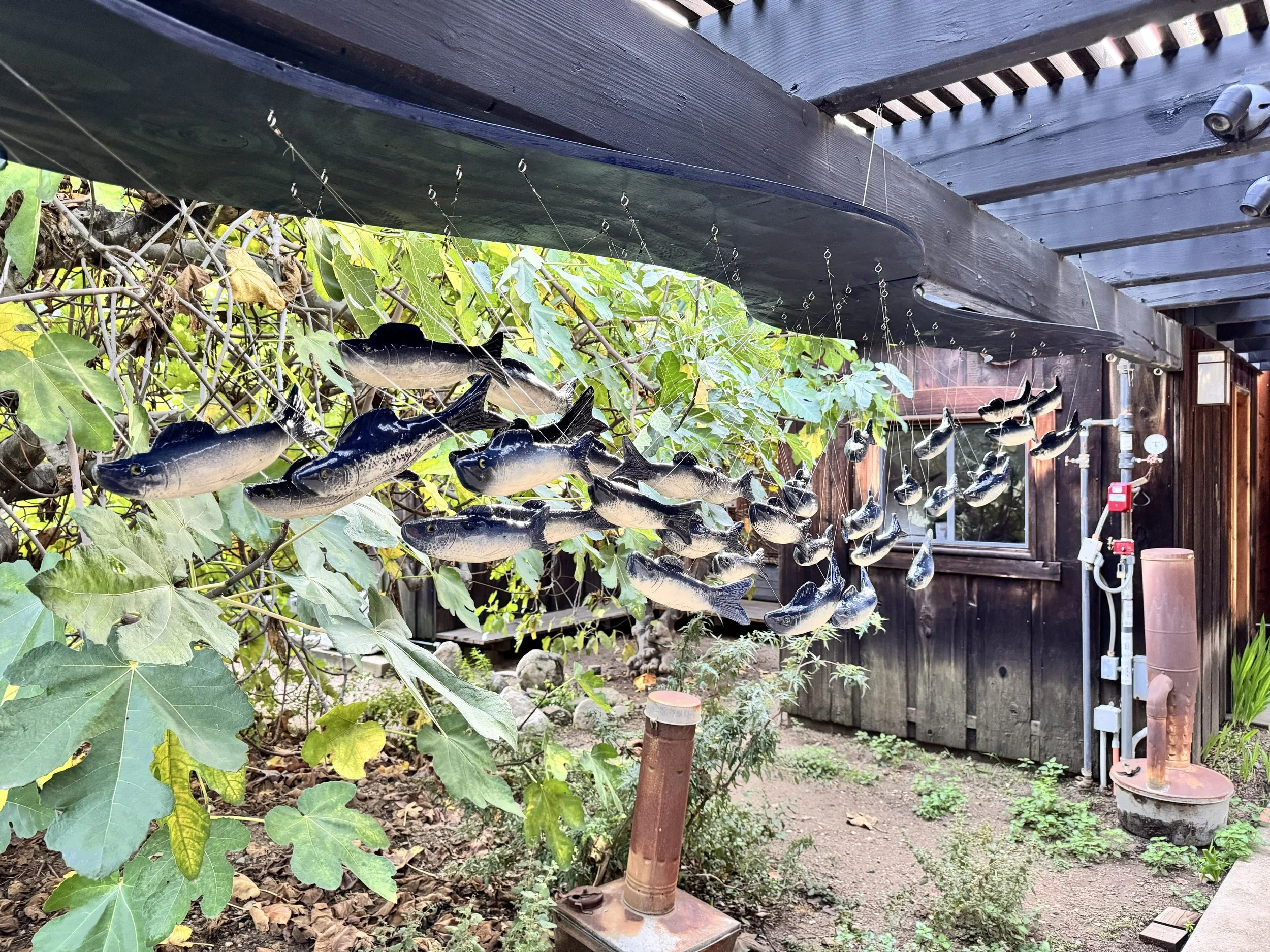 Fishing lure sculptures resembling small fish hanging from a wooden structure in an outdoor garden setting.