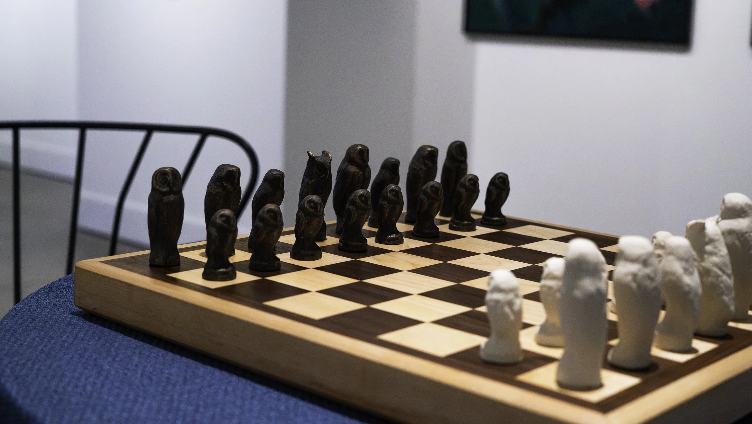 A chessboard set up for a game with black and white pieces, including pawns, on a wooden board in a room with a white wall and a black chair.