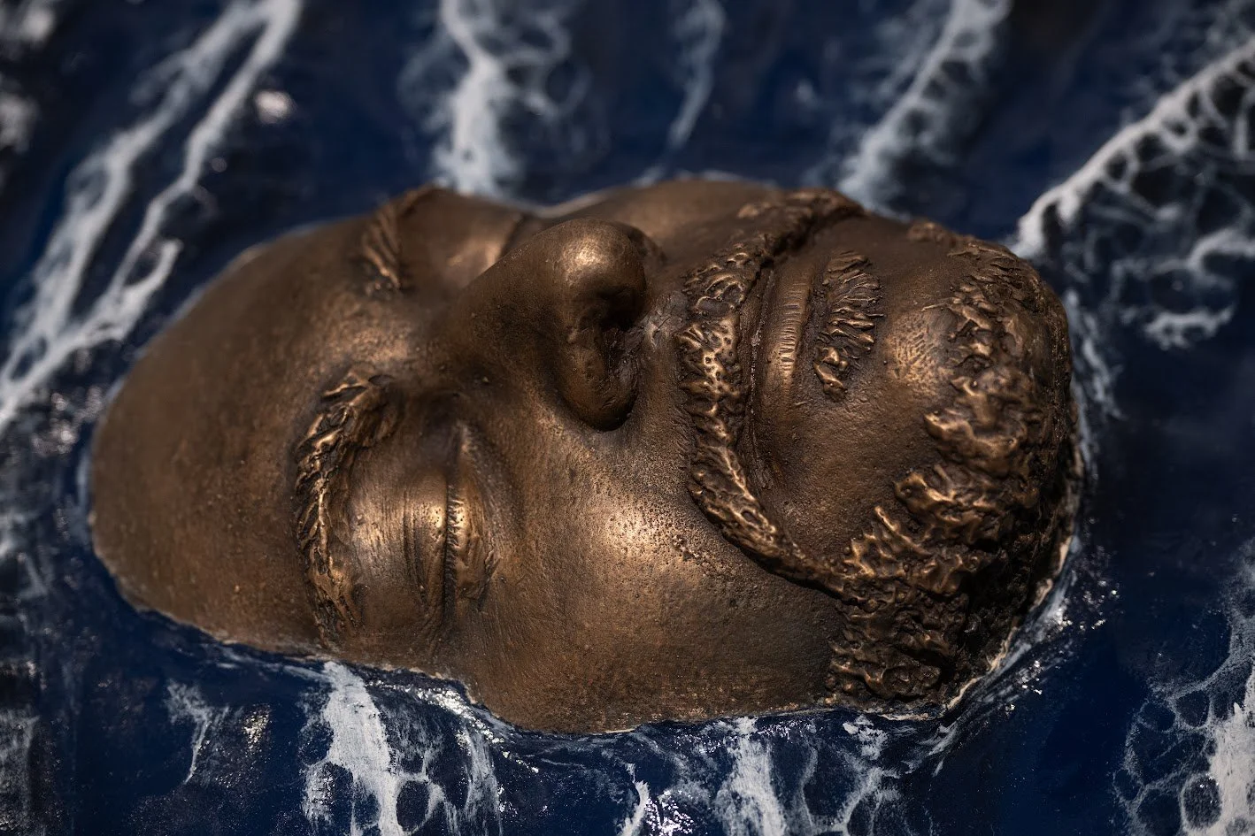 Close-up of a bronze statue of a person's face with eyes closed, partly submerged in water.