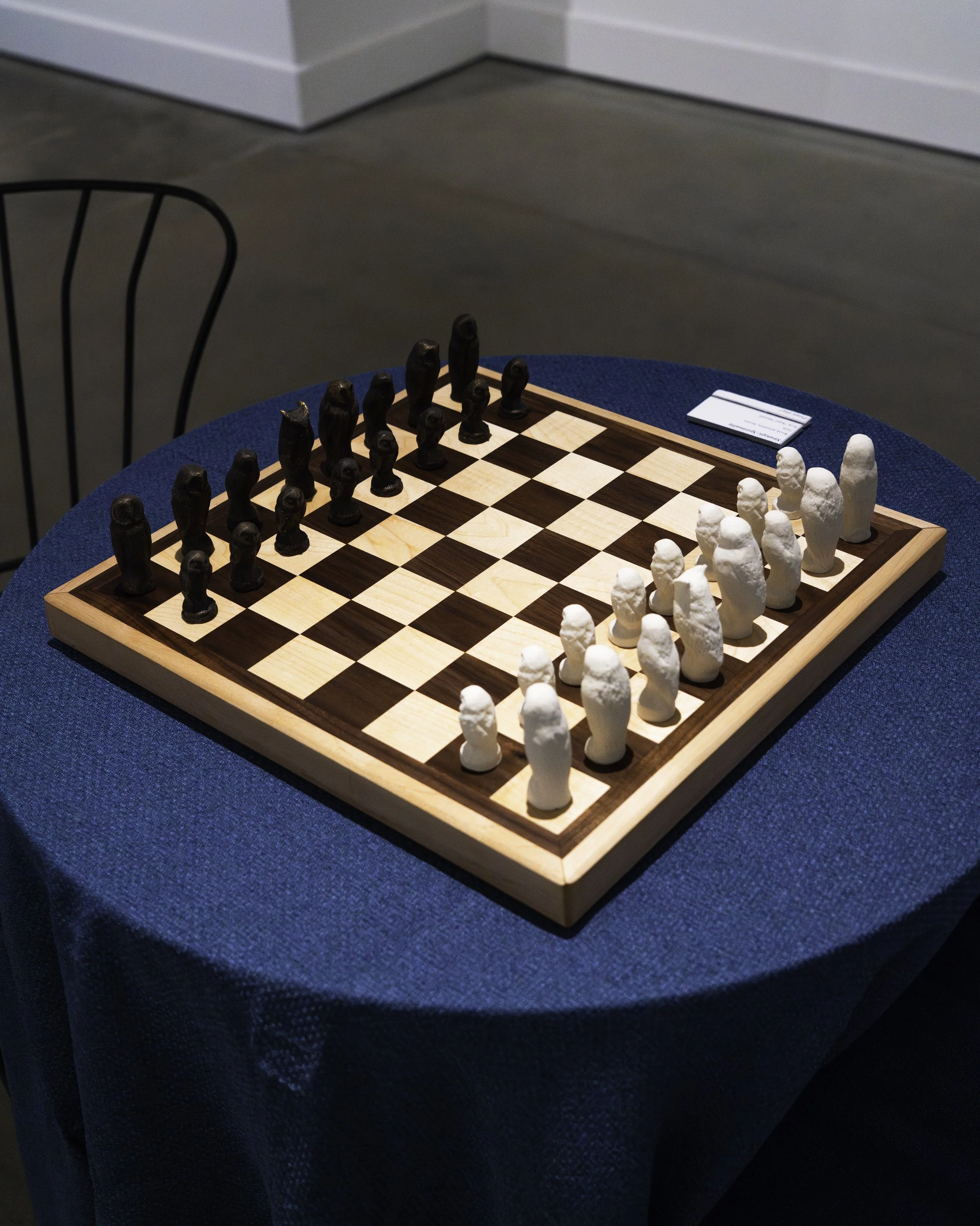 A chessboard with black and white chess pieces set up for a game, placed on a round blue table, with a chair nearby in a room.