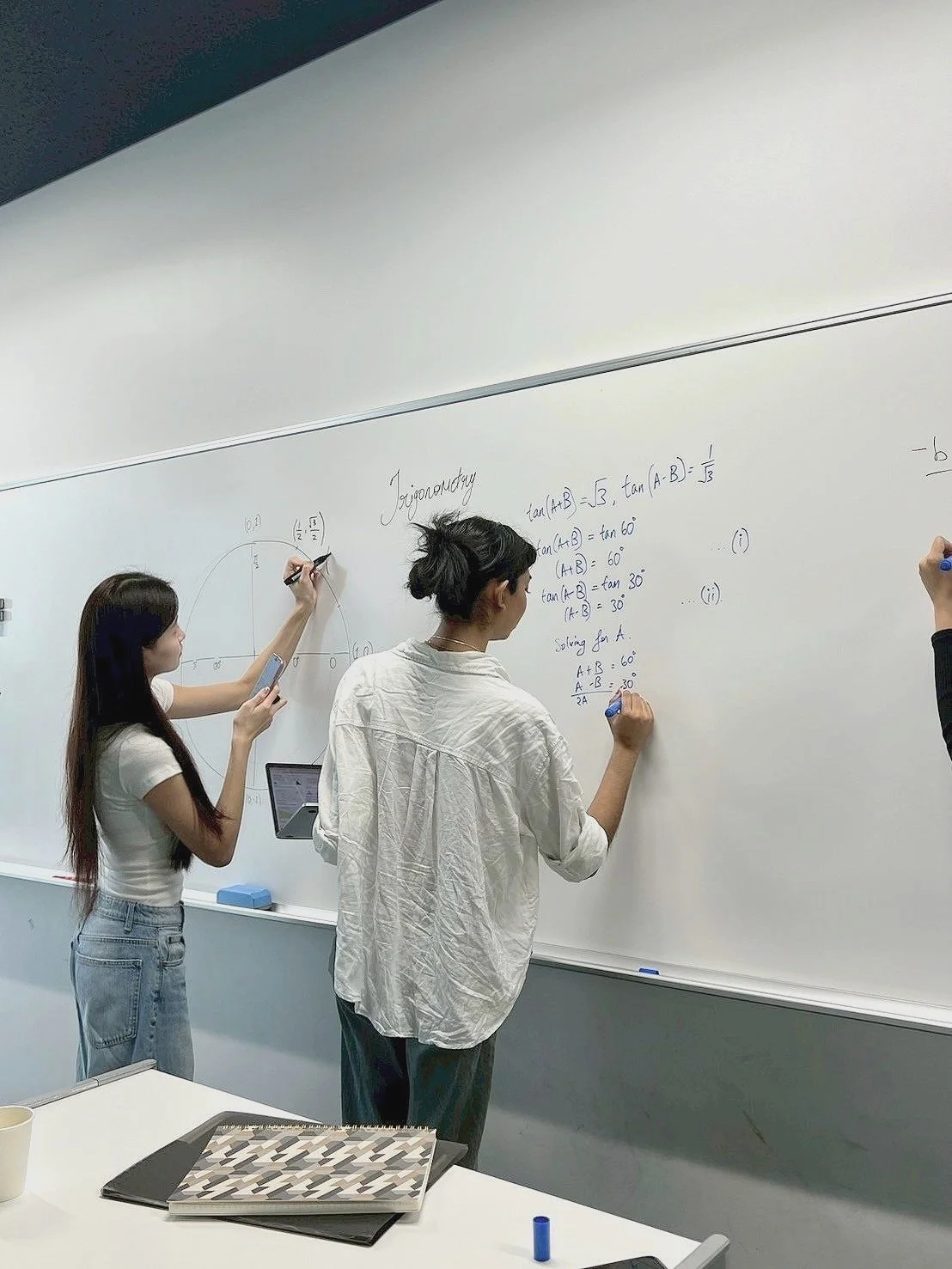 Three students writing on a classroom whiteboard filled with mathematical equations and diagrams.