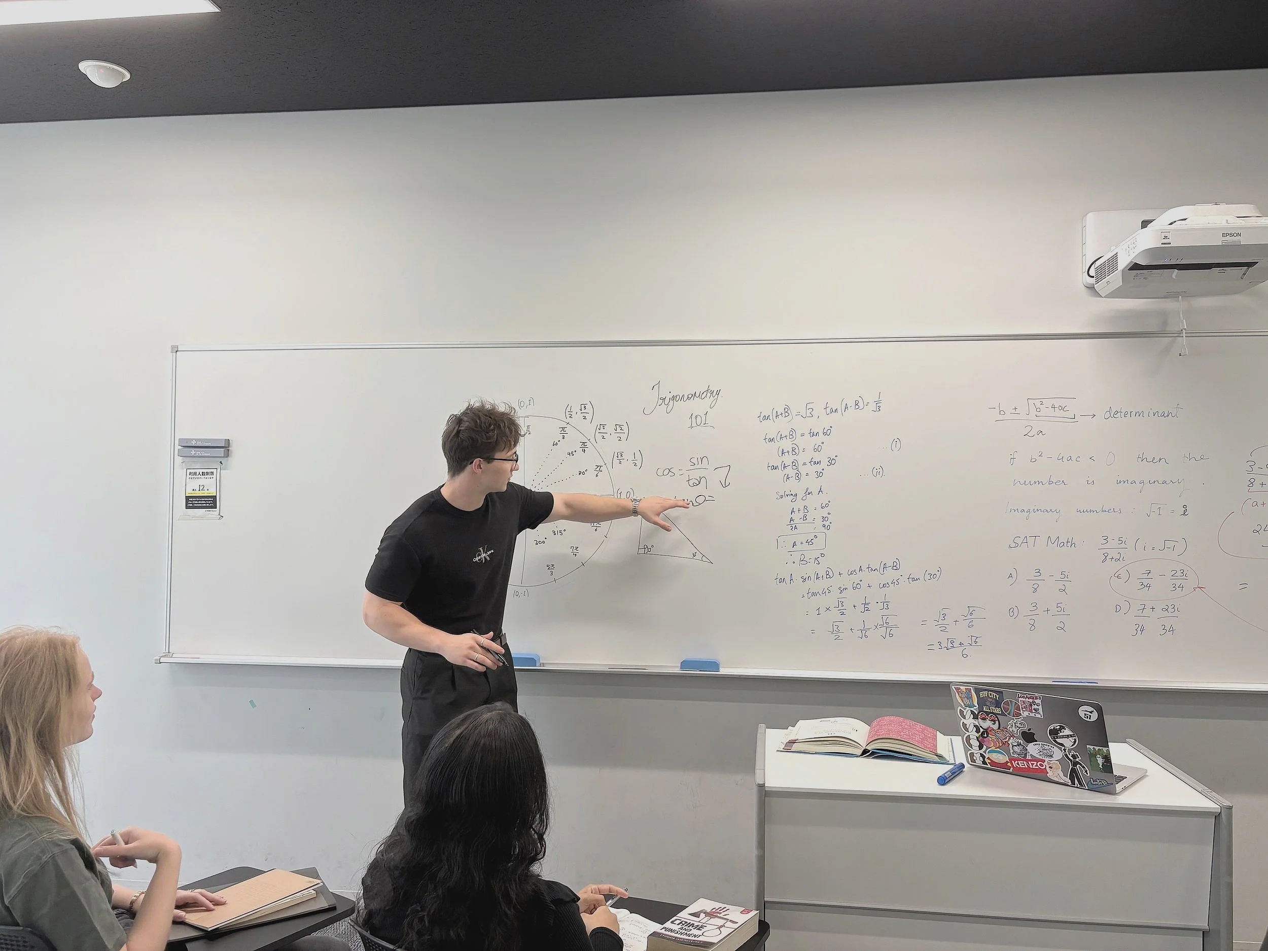 A college classroom with a male professor teaching mathematics on a whiteboard to two female students seated at desks.