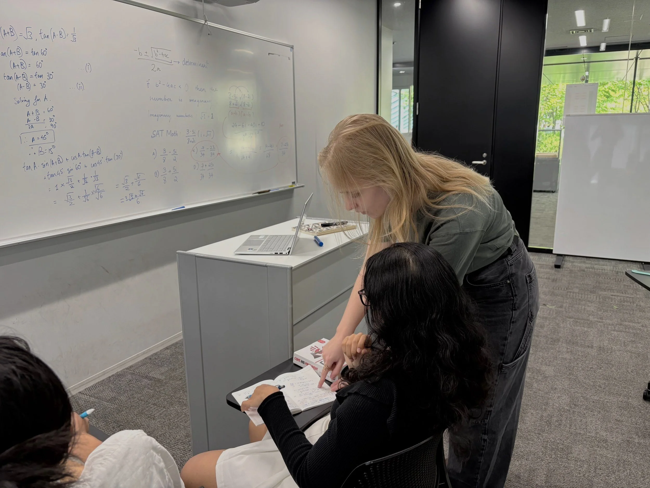 A teacher helping a student with her notes during class in a classroom with a whiteboard filled with mathematical equations.
