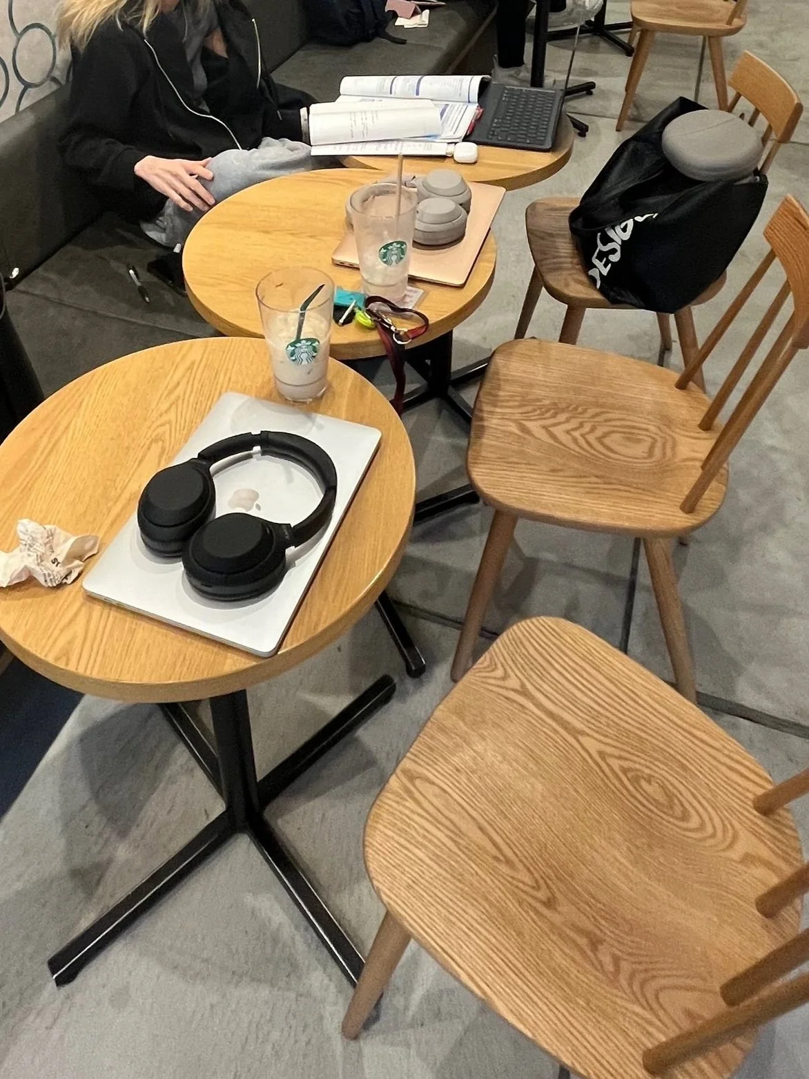A table with two Starbucks drinks, a pair of headphones, and some opened books, surrounded by additional chairs, with a person sitting in the background.