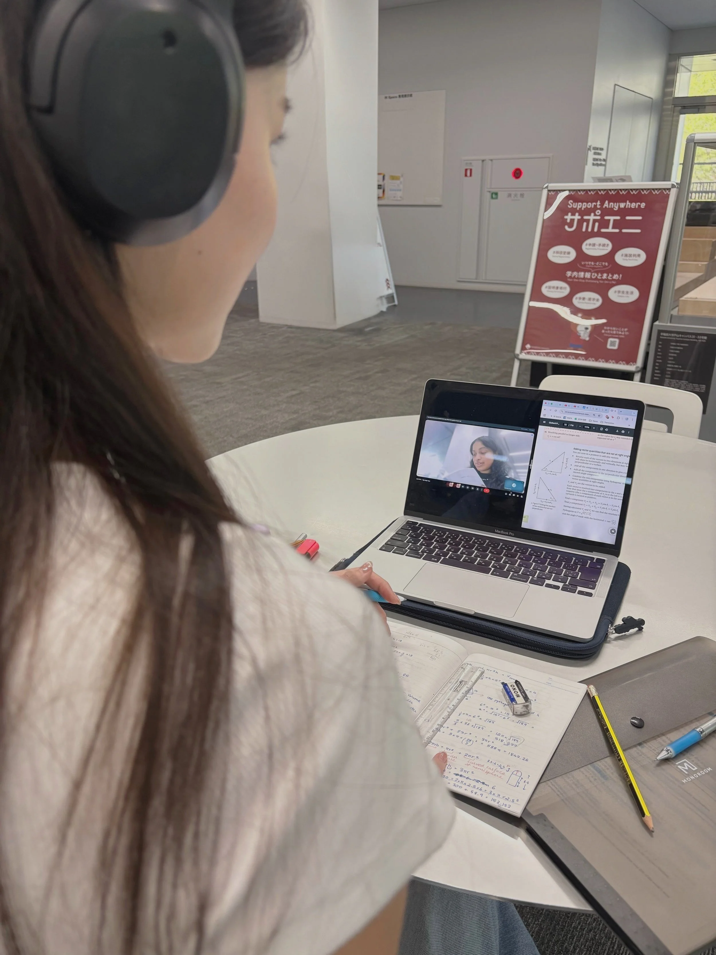 A woman wearing headphones studying at a round table with open book, notebook, stylus, laptop, and a calculator. She is attending an online class or meeting on her laptop.