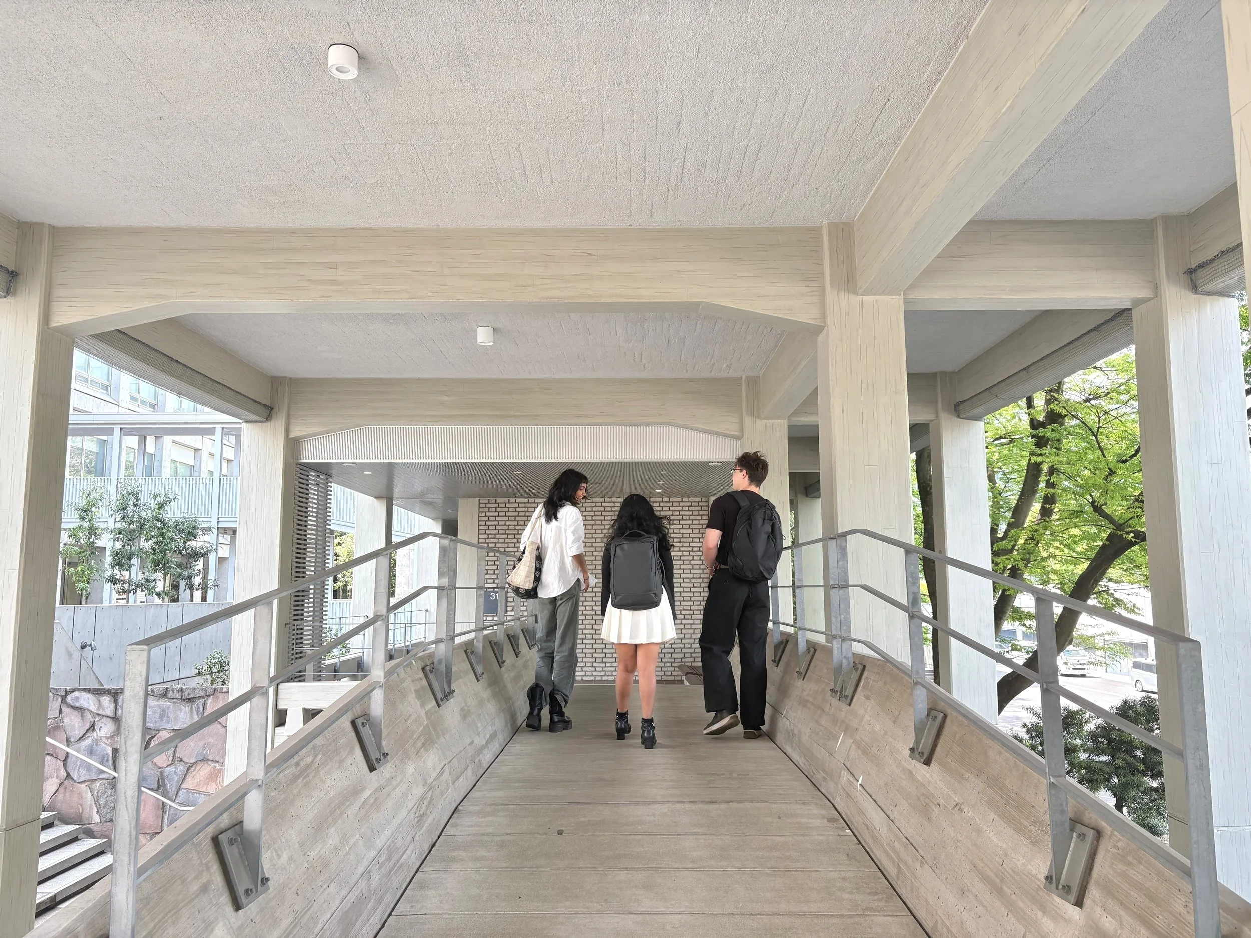 Three people walk up an indoor ramp with metal railings, inside a modern building with concrete pillars and large windows, with trees visible outside.