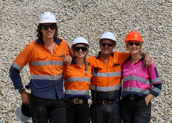 Four women wearing safety helmets and orange and pink safety shirts standing on rocky terrain, smiling with arms around each other.