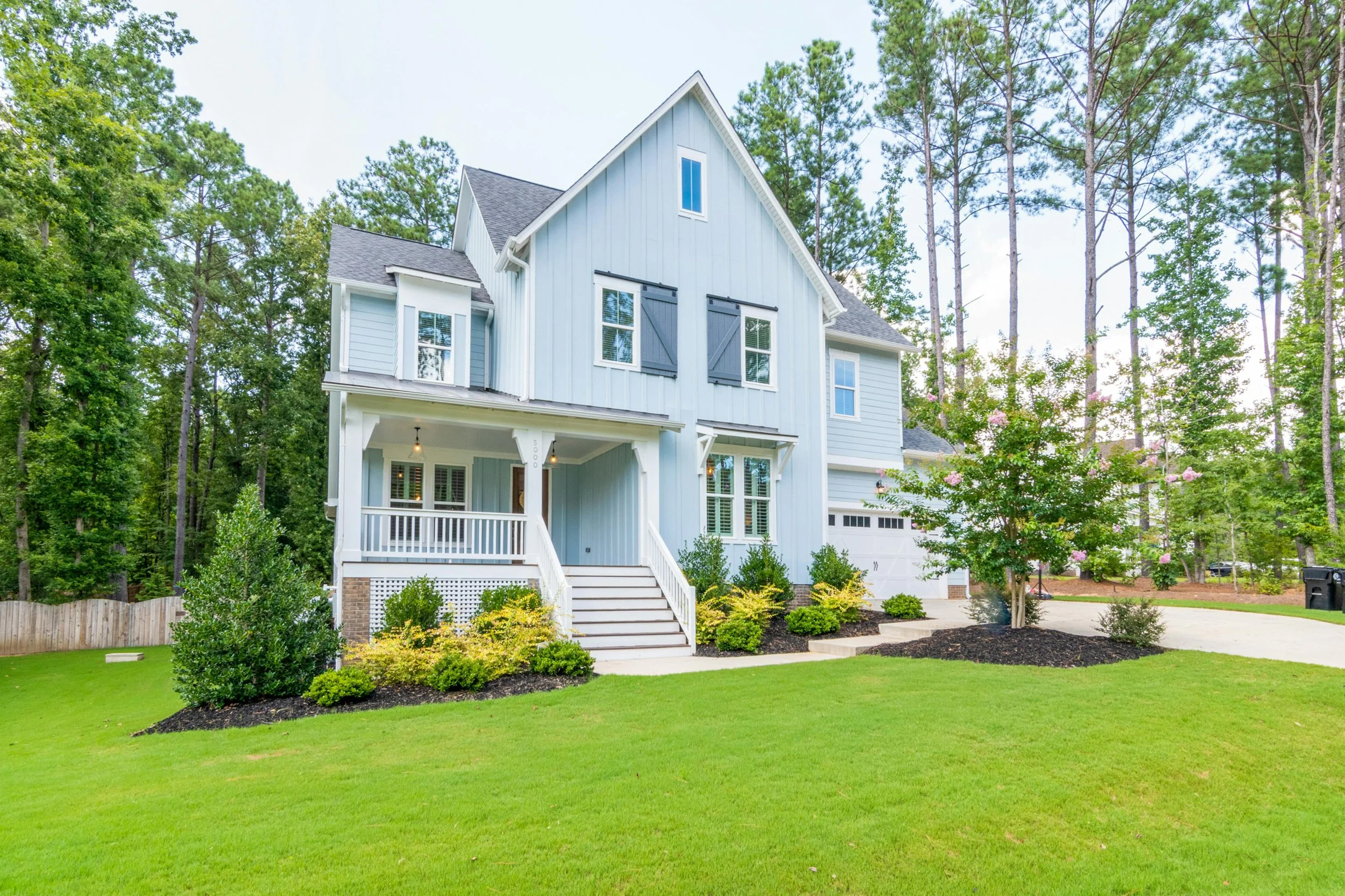 Light blue two-story house with a covered front porch, white railings, and stairs leading up to the entrance, surrounded by a green lawn, bushes, and tall trees.
