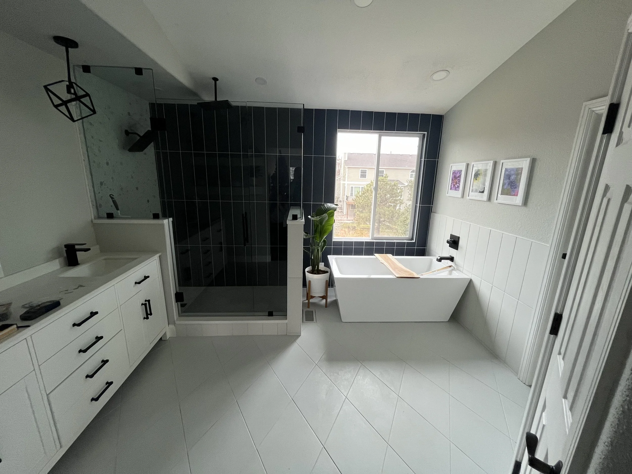Interior view of a modern bathroom with a window, white vanity with black handles, a black-tiled shower enclosure, and a bathtub with a wooden tray, decorated with framed floral artwork on the wall.