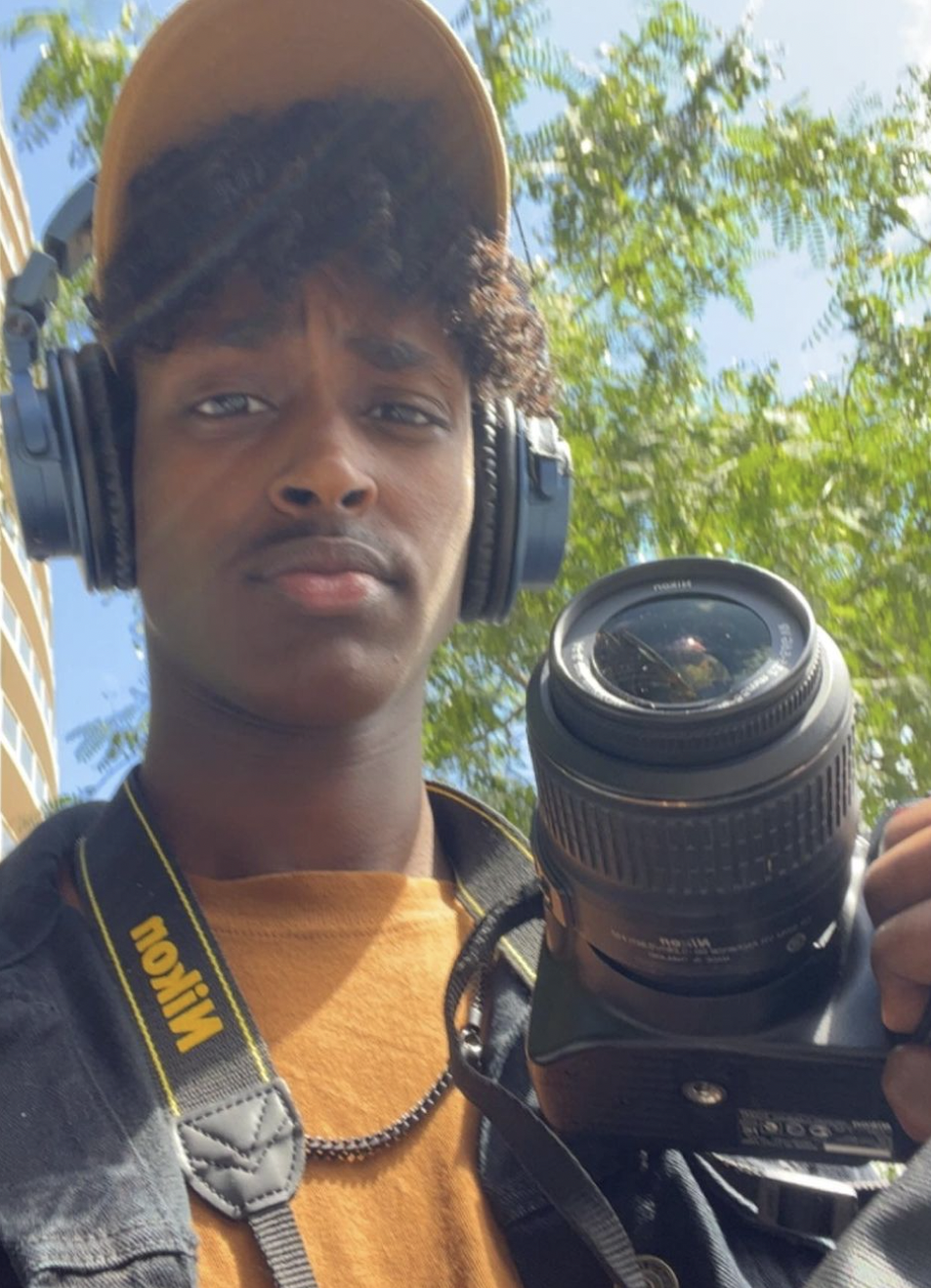 Young man taking a selfie outdoors with a Nikon camera, headphones, and a hat, standing under green trees and blue sky.