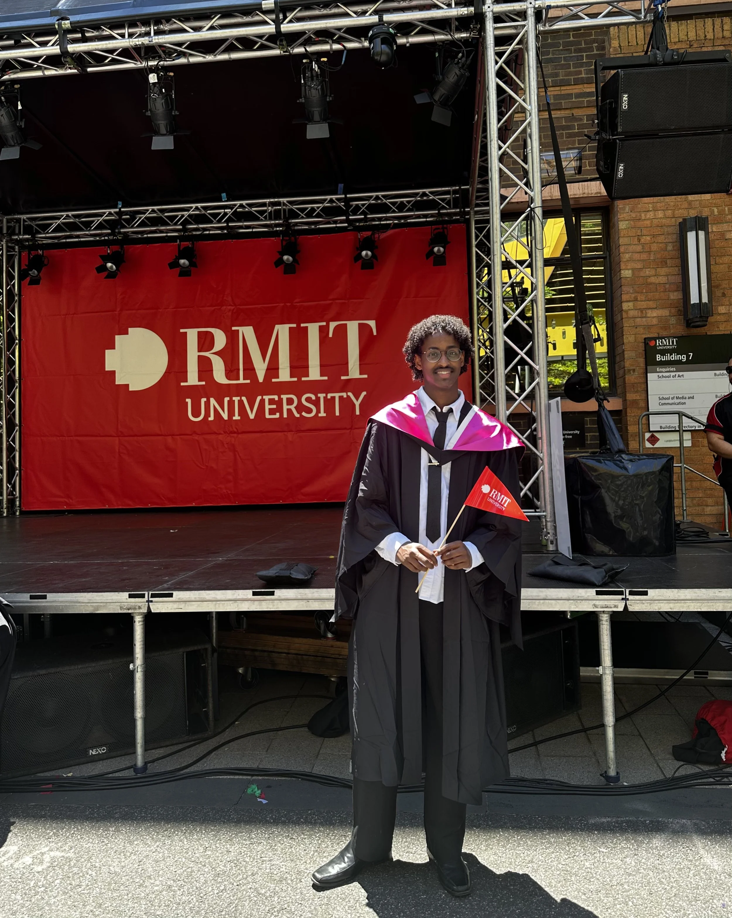 A young man wearing graduation gown and hood holding a red RMIT University flag standing outdoors in front of a stage with a red backdrop and speaker equipment, celebrating graduation.